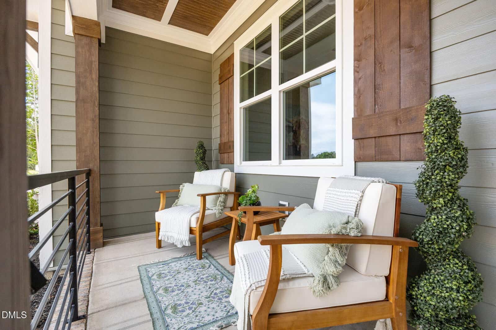Two cushioned wooden chairs with throw pillows and blankets sit on a cozy porch, accented by a rug and topiary plants, with large windows and wood shutters behind them.