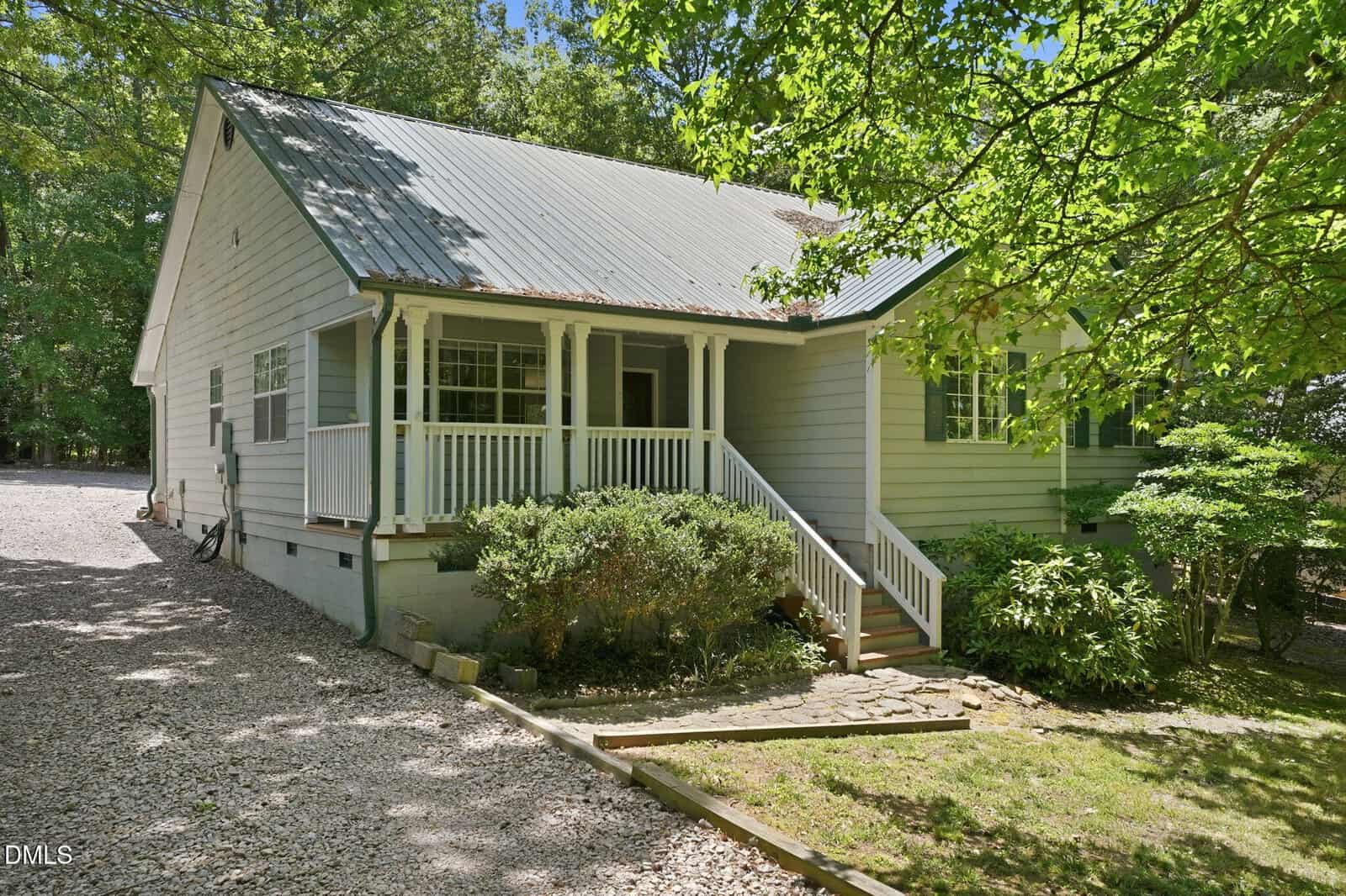 A light gray house with a metal roof, covered front porch, and white railings, surrounded by trees and bushes, with a gravel driveway along the side. Sunlight filters through the leafy branches above.