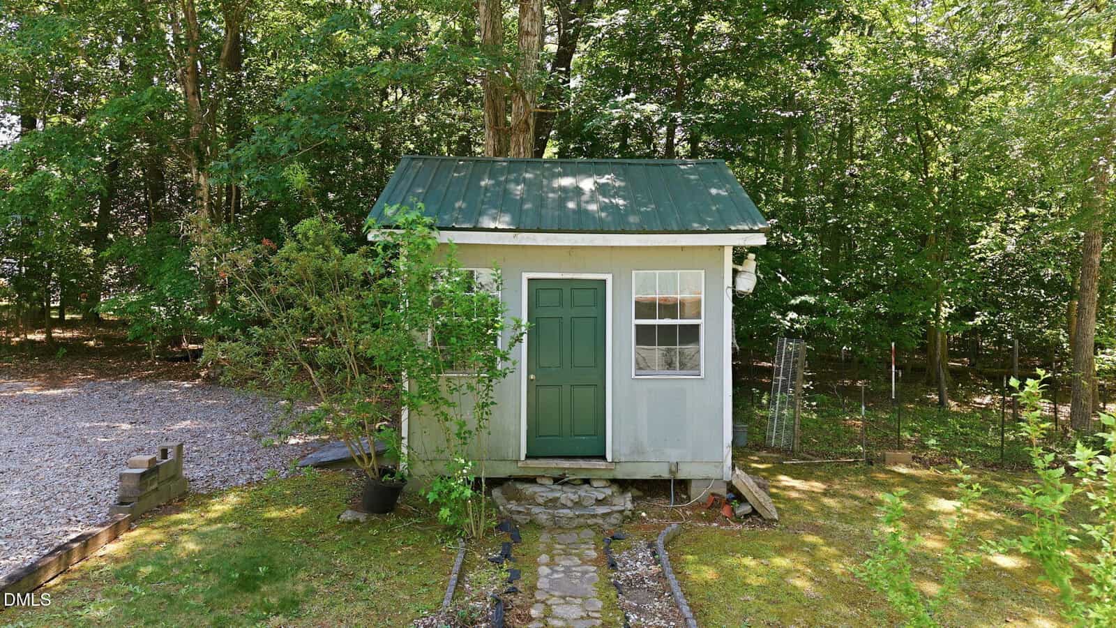 A small green-roofed shed with a green door and white trim sits in a shaded yard, surrounded by trees and plants, with a stone path leading to the entrance.