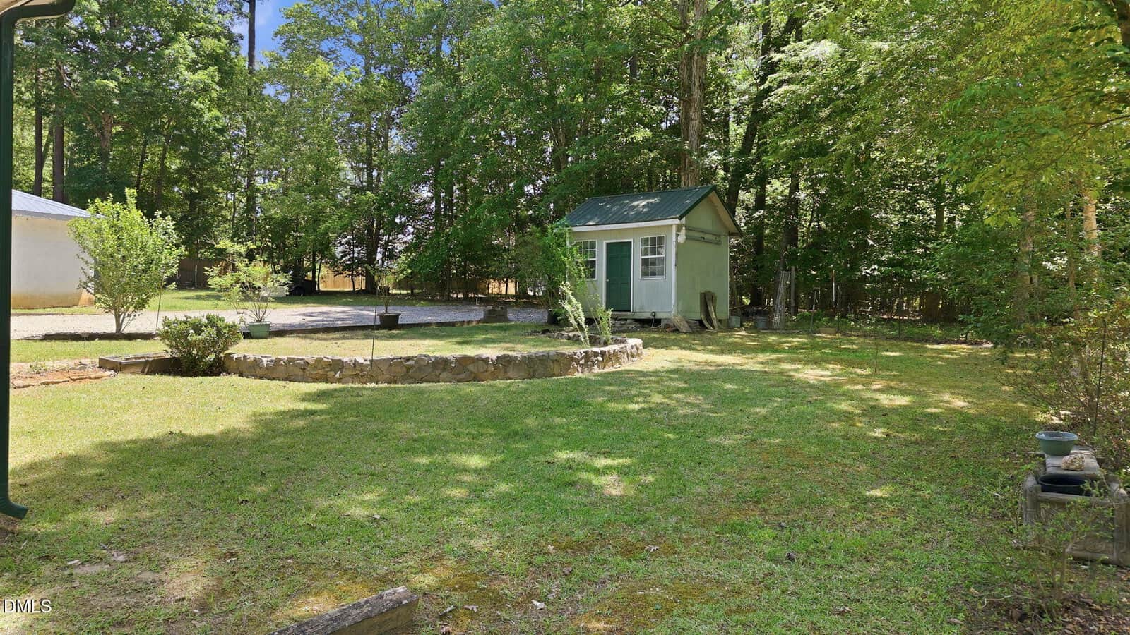 Sunny backyard with grass, a small stone fire pit, a green-roofed shed, and surrounding trees. Potted plants and a gravel area are also visible.