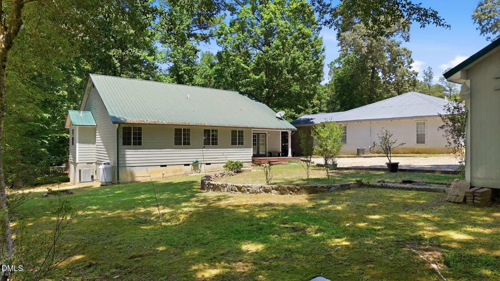 A light gray house with a green metal roof sits among trees. There’s a small deck in back, a shed on the right, and a grassy yard with a circular stone-bordered garden bed in the center.