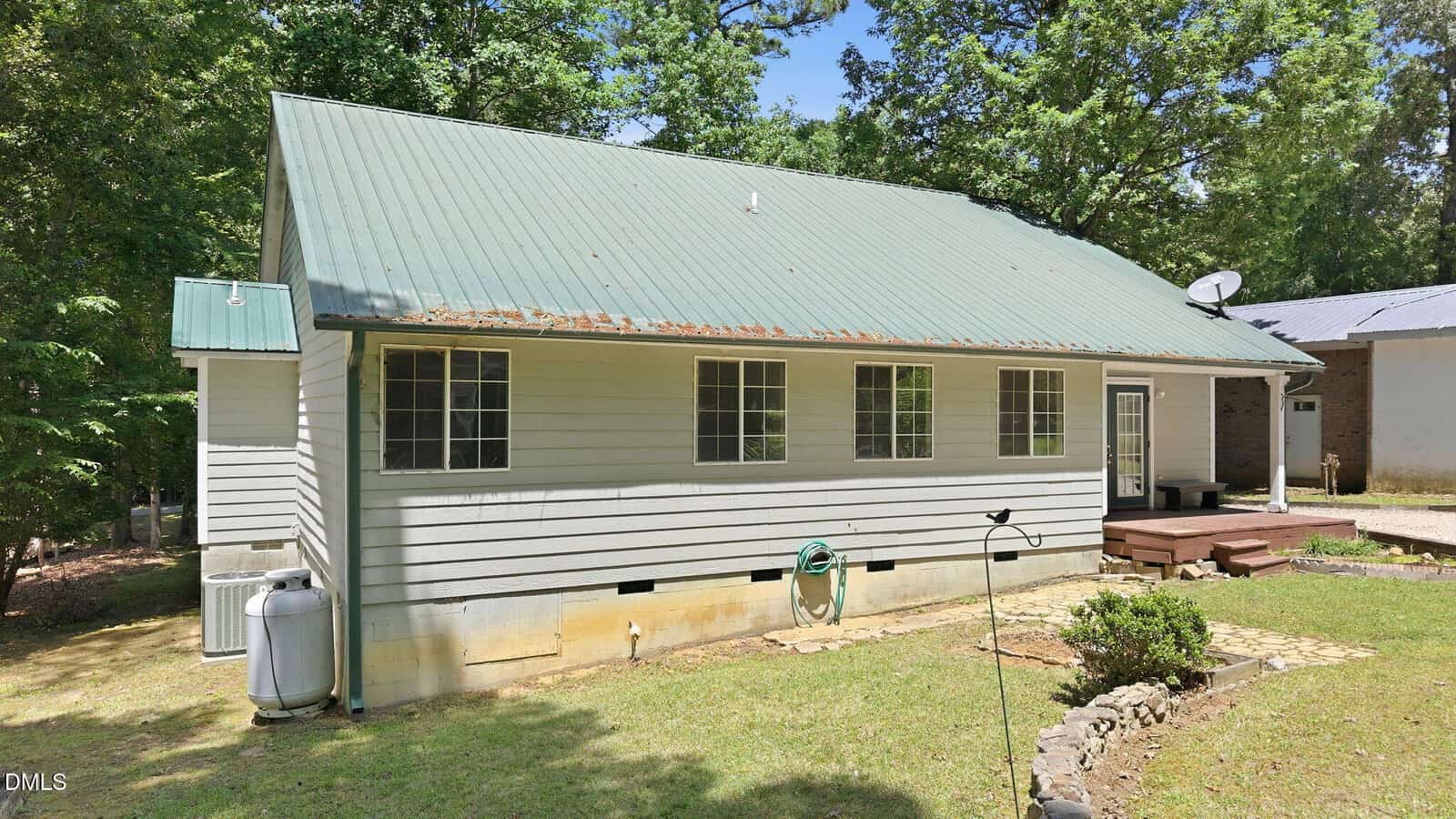 A single-story house with light gray siding, a green metal roof, several windows, a wood deck, and a propane tank on a grassy yard bordered by trees.