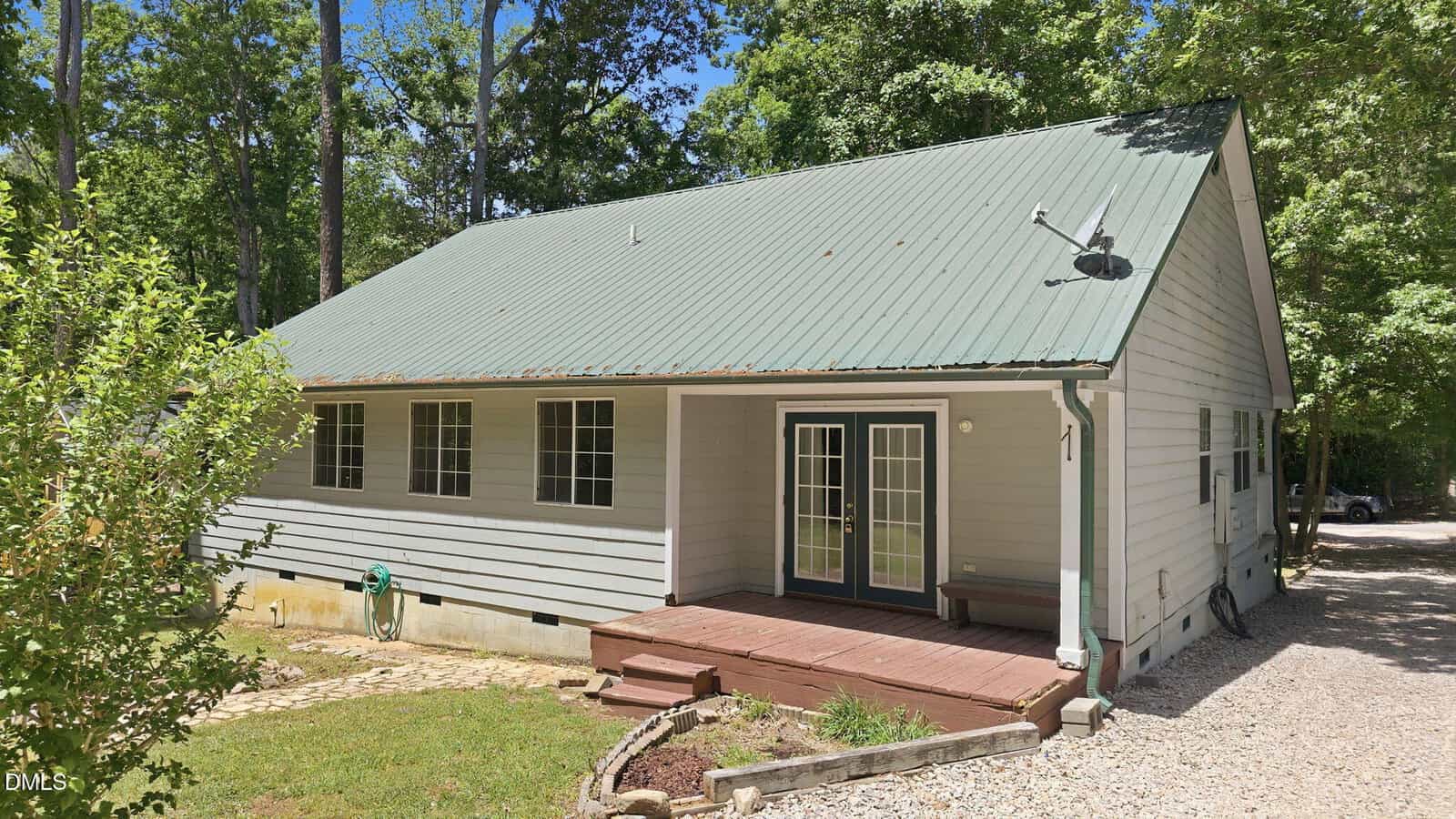 A small white house with a green metal roof, wooden front porch, and French doors. The house is surrounded by trees, grass, and a gravel driveway on the right side.