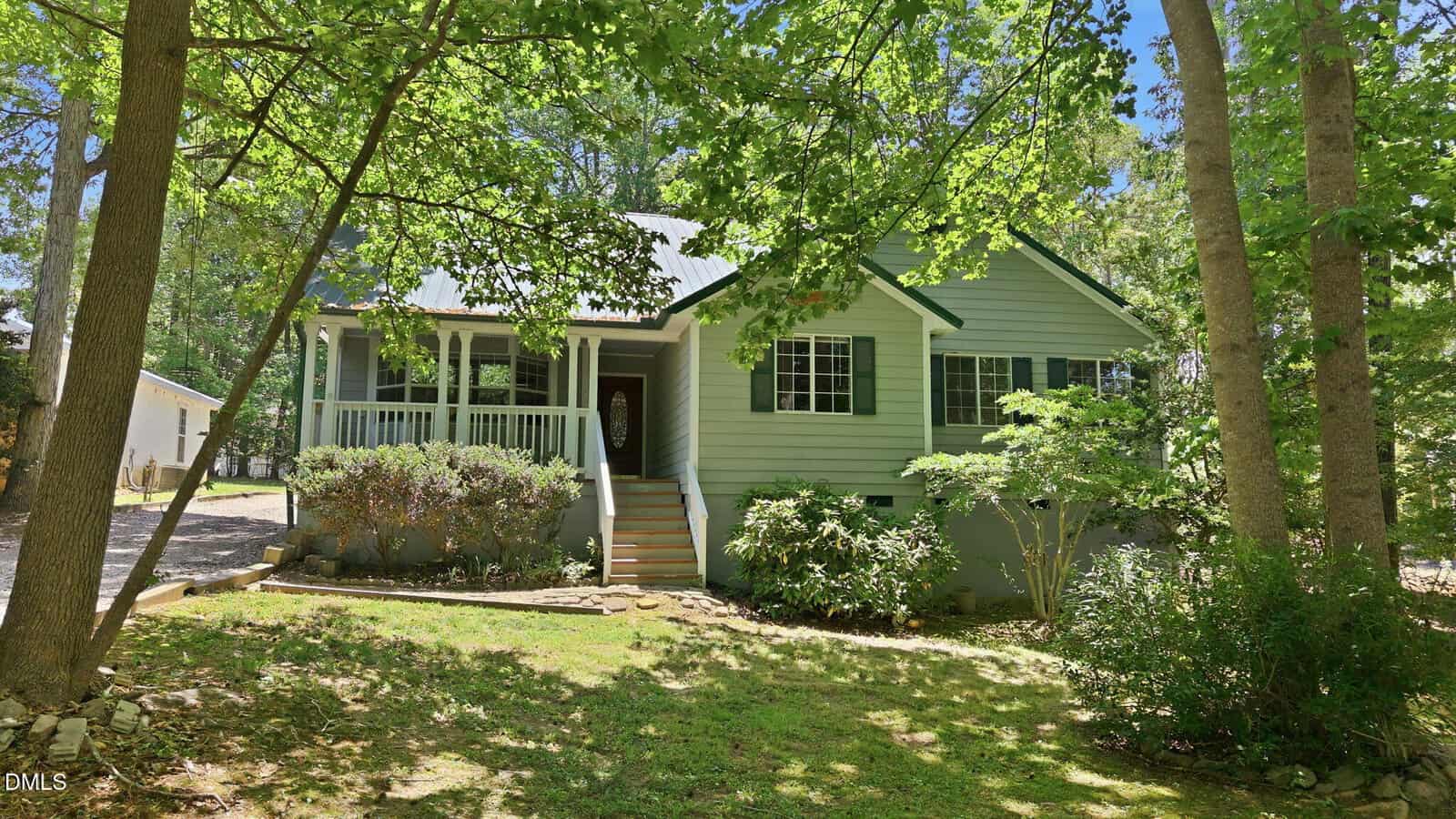 A light green house with a front porch, white railings, and steps, surrounded by trees and shrubs, with sunlight filtering through the leaves onto the lawn.