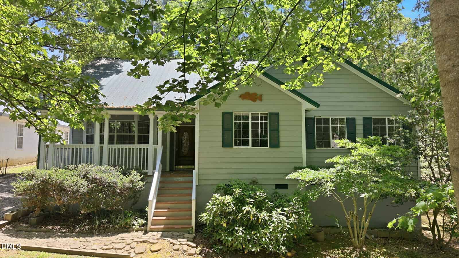 Light green cottage-style house with a metal roof, front porch, and green shutters, surrounded by leafy trees and bushes in bright sunlight. Steps lead to the front door, and a fish decoration is above the windows.