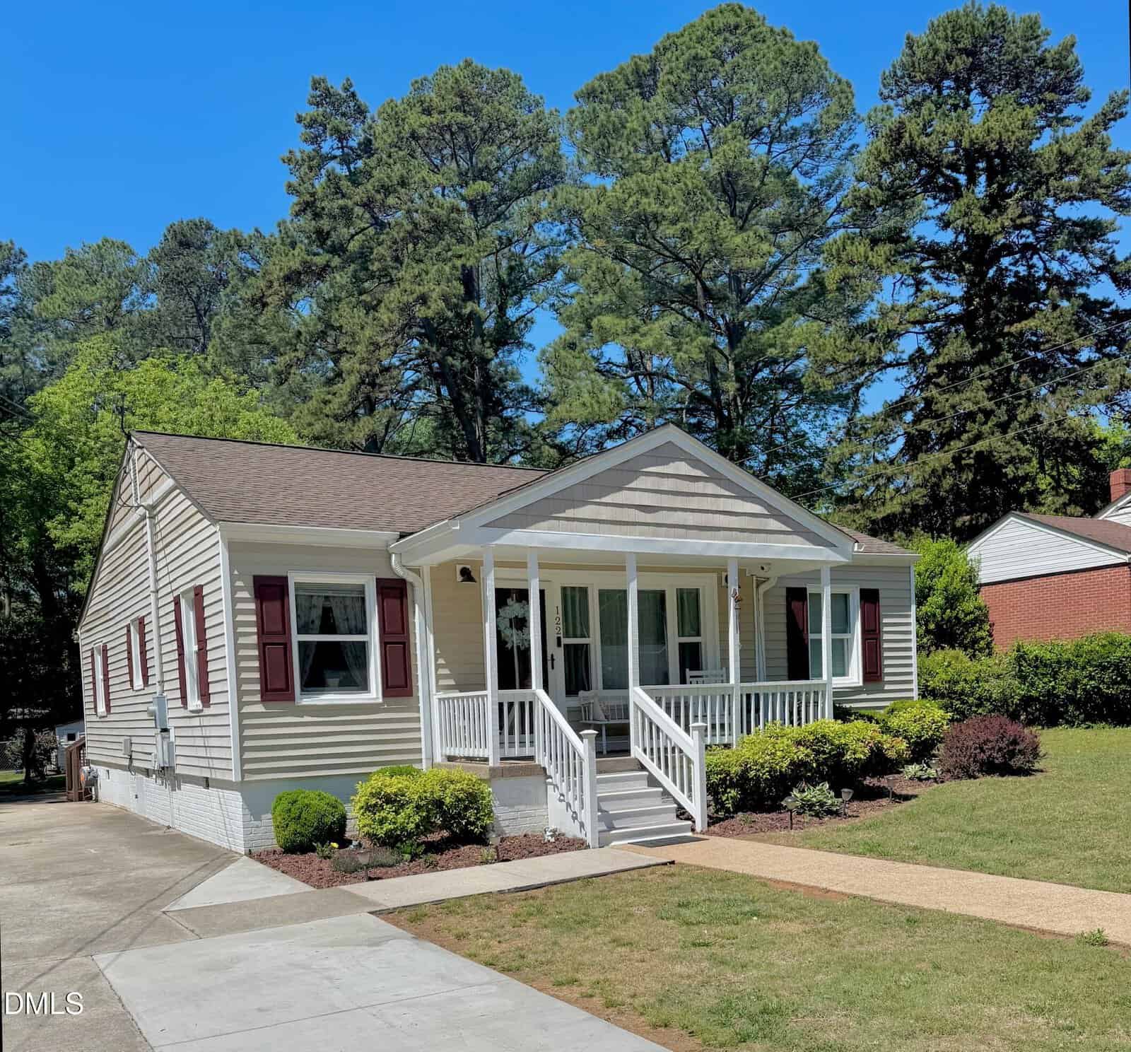 Single-story beige house with white trim and maroon shutters, a front porch with white railings, and a well-kept lawn with shrubs. Tall trees and a driveway are visible in the background under a clear blue sky.