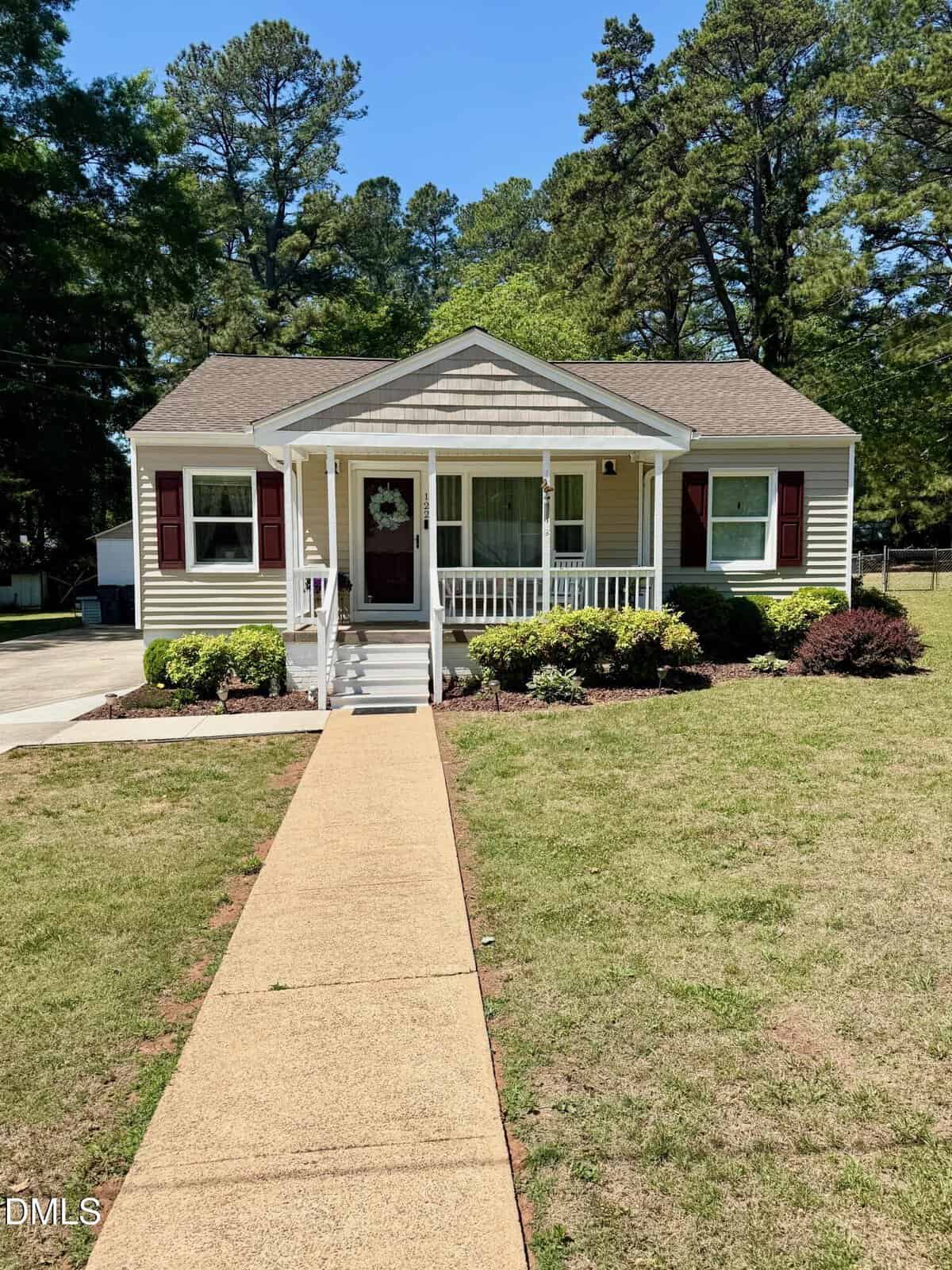 A small, single-story yellow house with white trim and a covered front porch. Bushes line the front of the house, and a concrete walkway leads from the sidewalk to the porch. Tall trees are in the background.