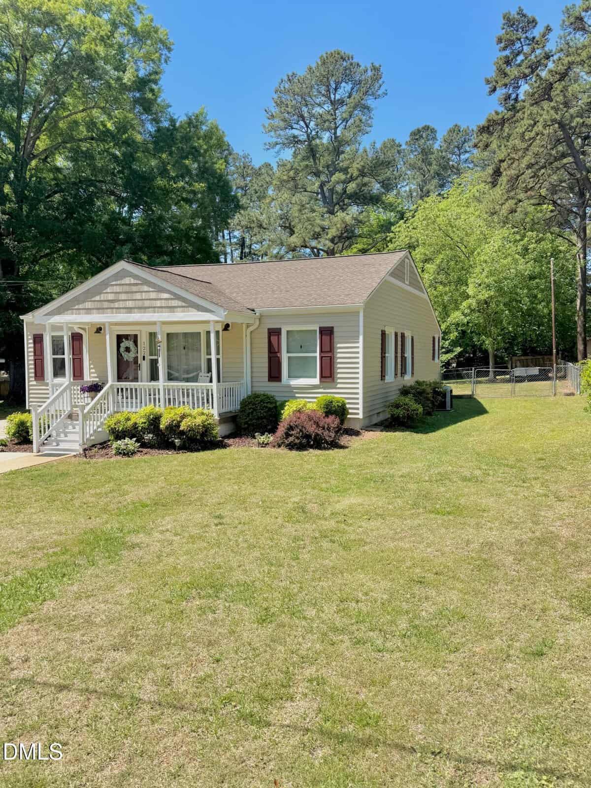 A small, single-story yellow house with burgundy shutters and a covered front porch sits on a large grassy lawn, surrounded by trees and bushes on a sunny day.