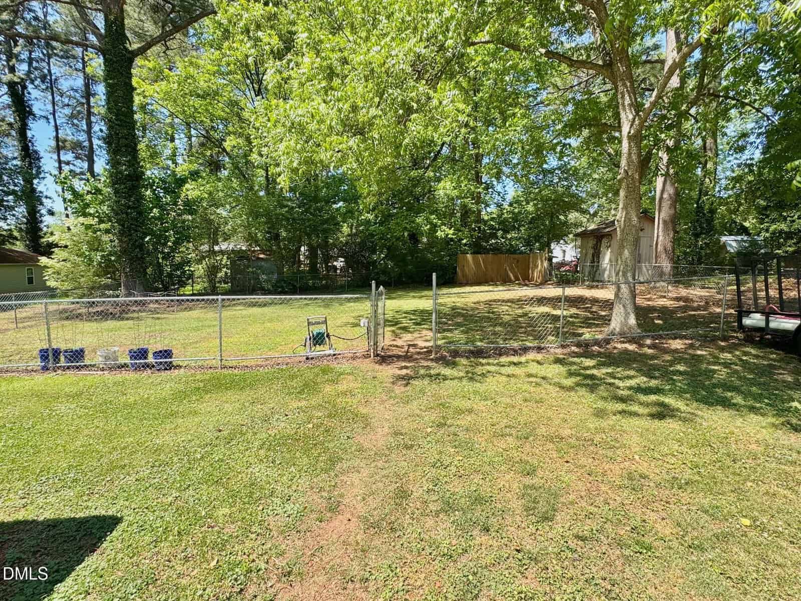 A fenced backyard with green grass, trees providing shade, and a small shed in the background. Sunlight filters through the leaves and there are some items near the fence.