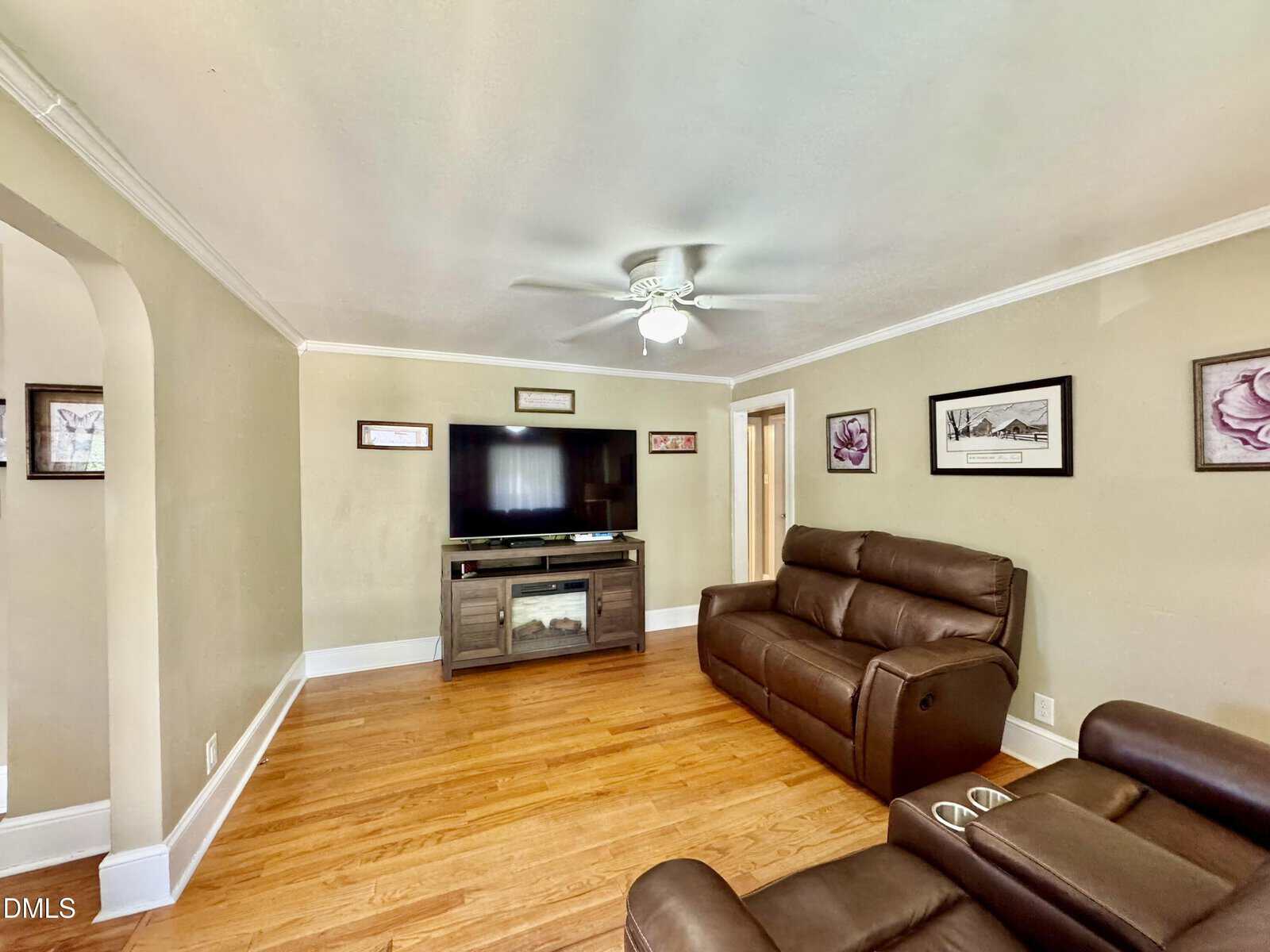A cozy living room with light brown walls, wood floors, a ceiling fan, brown leather sofas, a TV on a stand, and framed art on the walls. An arched doorway opens into another room.