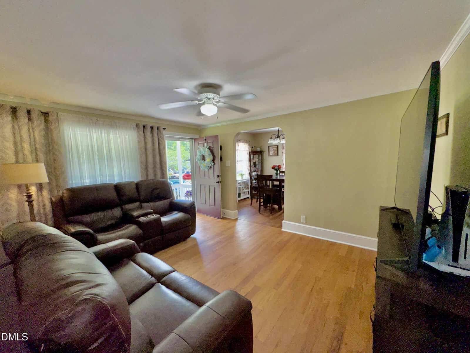 Living room with brown leather sofas, hardwood floors, TV on a stand, and ceiling fan. An open doorway leads to a dining area and the front door is open to the outside. Light from windows brightens the space.