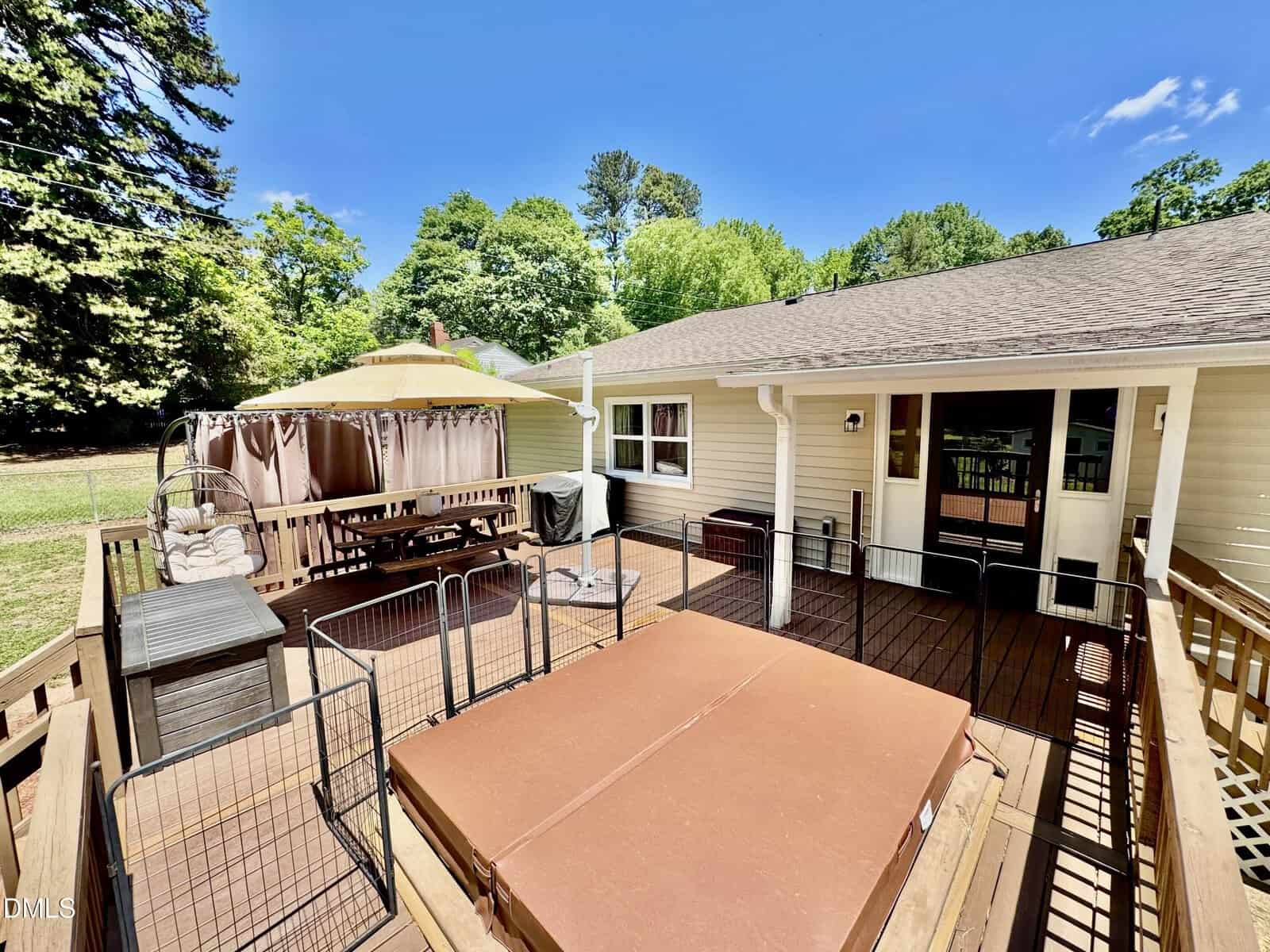 Spacious wooden backyard deck with a hot tub, outdoor dining table under a canopy, hanging chair, and seating area. The deck is attached to a light-colored house and surrounded by greenery under a blue sky.