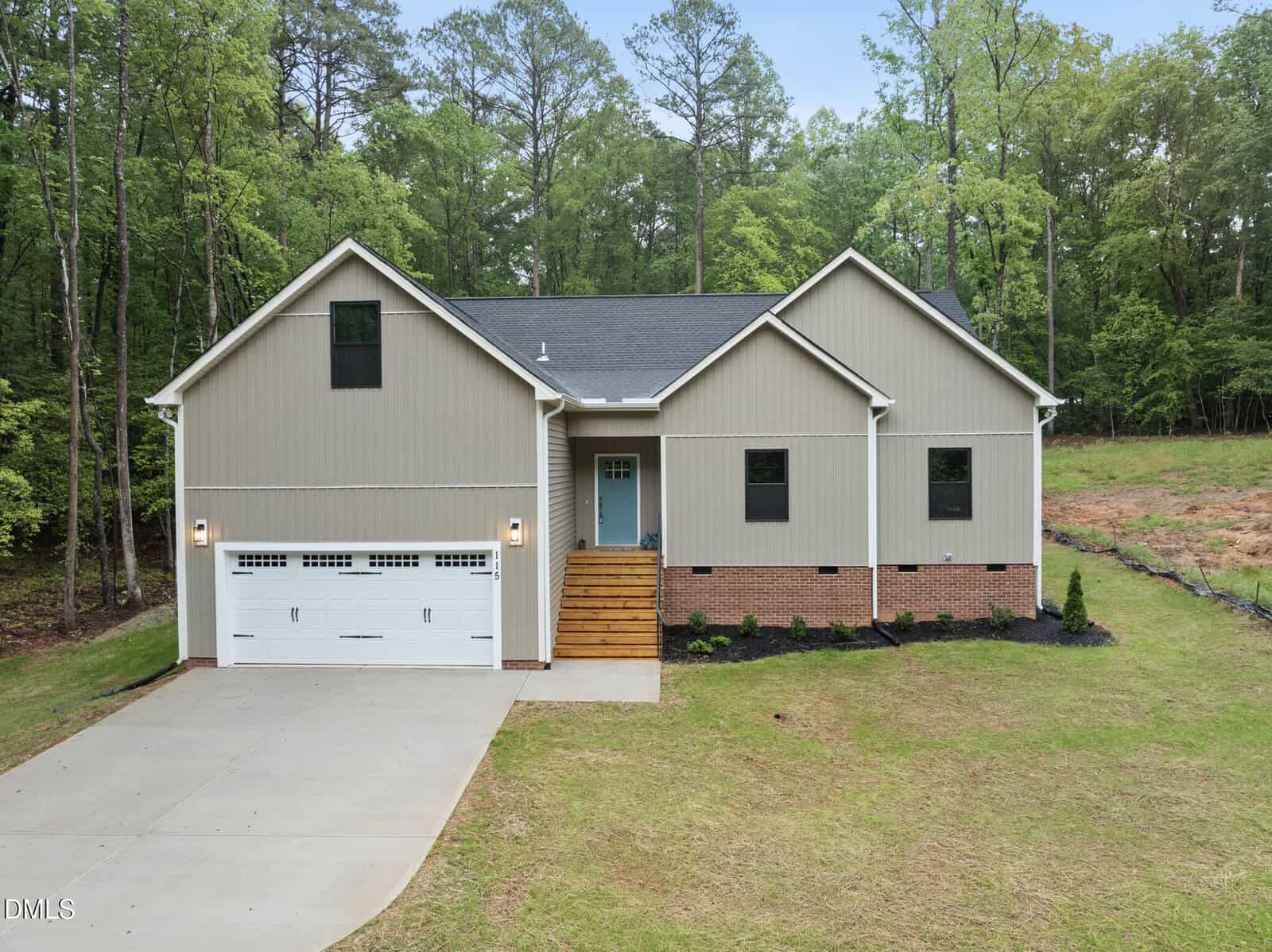 A modern single-story house with beige siding and brick accents, a two-car garage, and a wooden front porch, set against a backdrop of tall green trees and a spacious yard.