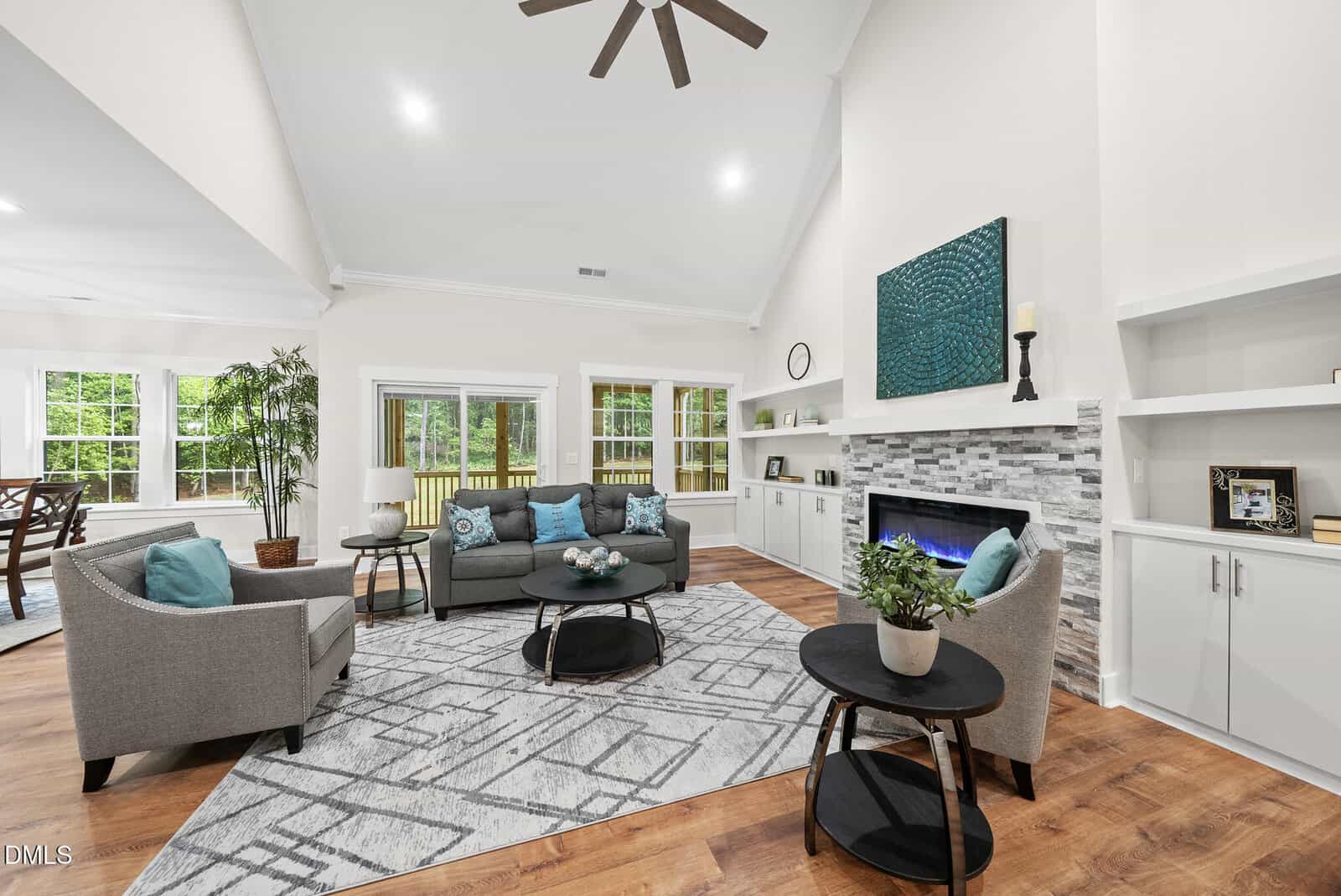 A bright, modern living room with two gray sofas, blue accents, a patterned rug, fireplace with stone surround, built-in shelving, wood floors, and large windows overlooking a green backyard.