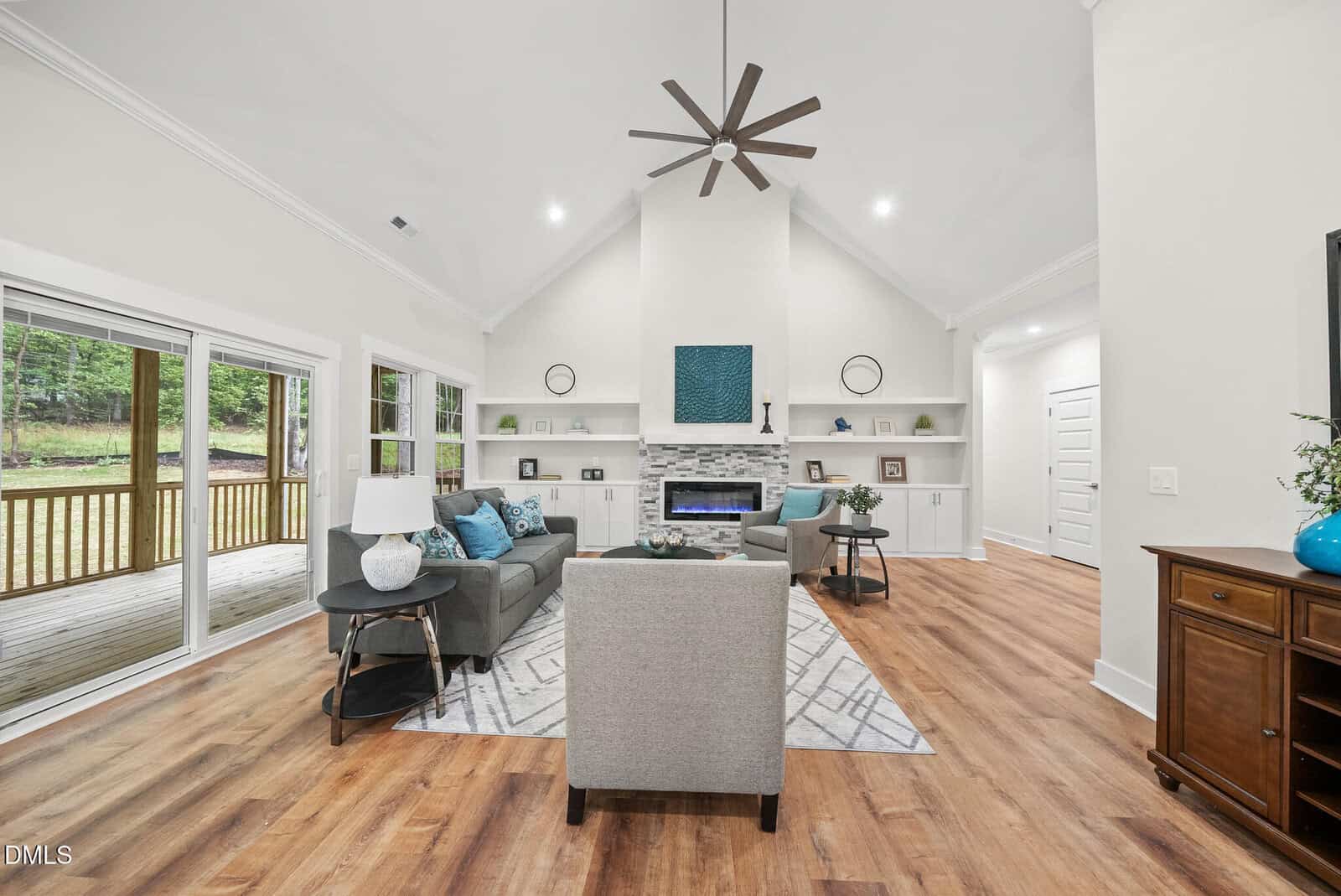 Modern living room with vaulted ceiling, light walls, wood floors, gray and blue furniture, fireplace with built-in shelves, large windows, glass doors to a deck, and a ceiling fan.