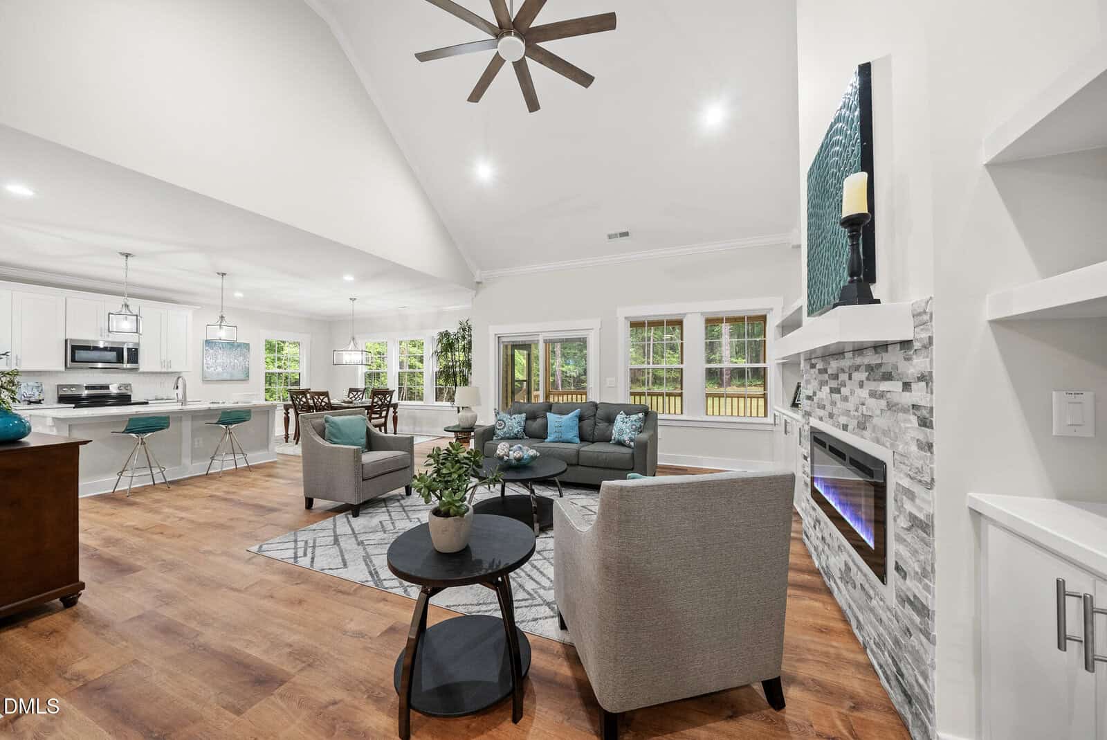 Modern open-concept living room with gray sofas, armchairs, fireplace, and kitchen area. Hardwood floors, large windows with outside view, and teal decor accents throughout.