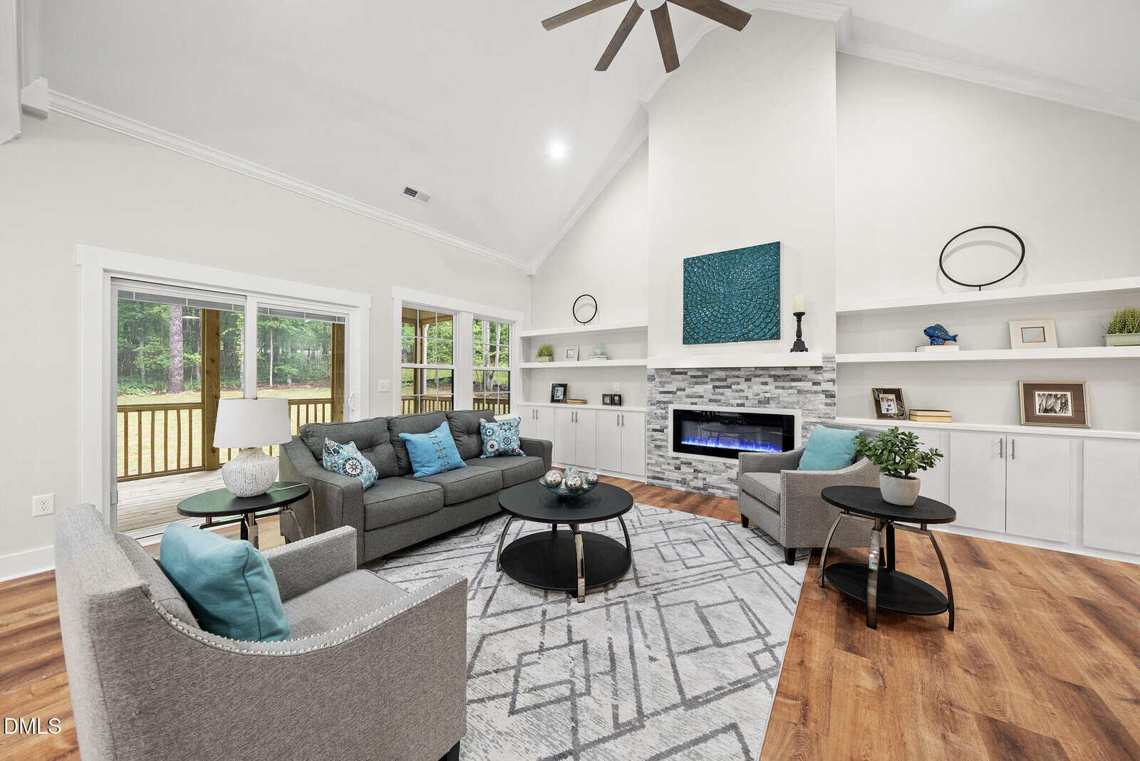 A modern living room with high ceilings, gray sofas, blue accents, a patterned rug, wall-mounted shelves, a fireplace, wood floors, and large windows overlooking a deck and greenery outside.