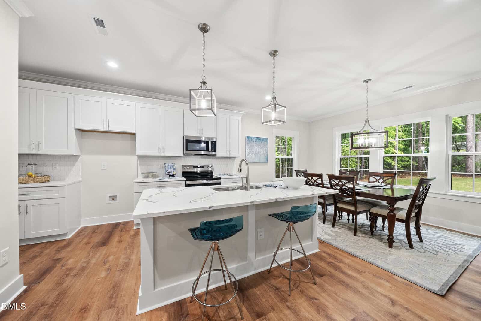 Modern kitchen with white cabinets, marble island with two teal bar stools, stainless steel appliances, and a dining area with a wooden table and six chairs by large windows overlooking a green backyard.