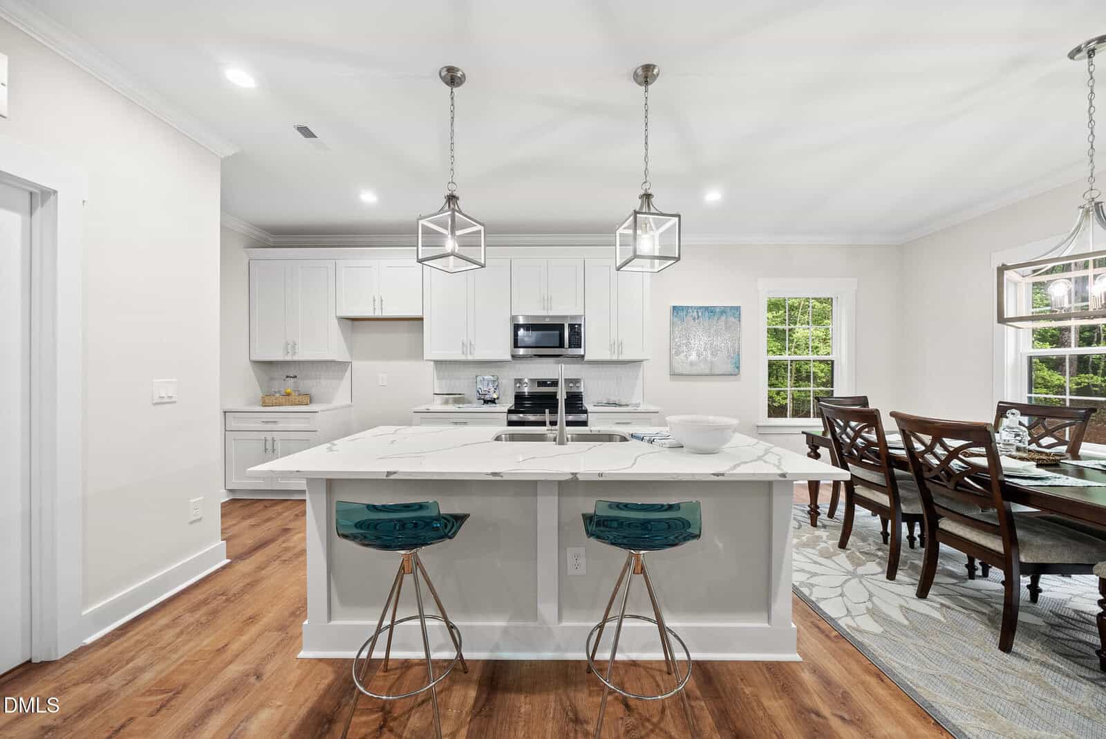 Modern kitchen with white cabinets, marble island, two teal barstools, pendant lights, stainless steel appliances, and a dining area with a wooden table and chairs near large windows letting in natural light.