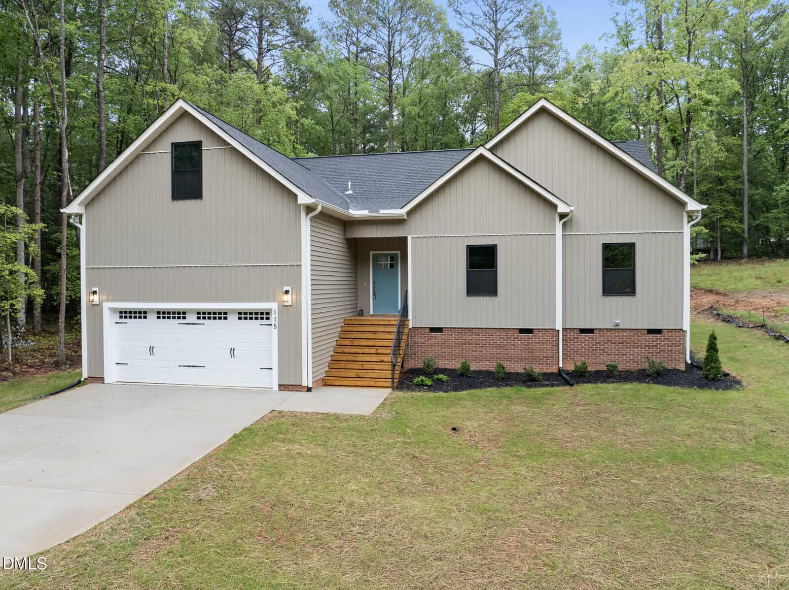 Modern single-story house with beige siding, brick foundation, a double garage, wooden steps leading to a blue front door, and a neat lawn; surrounded by tall trees in the background.
