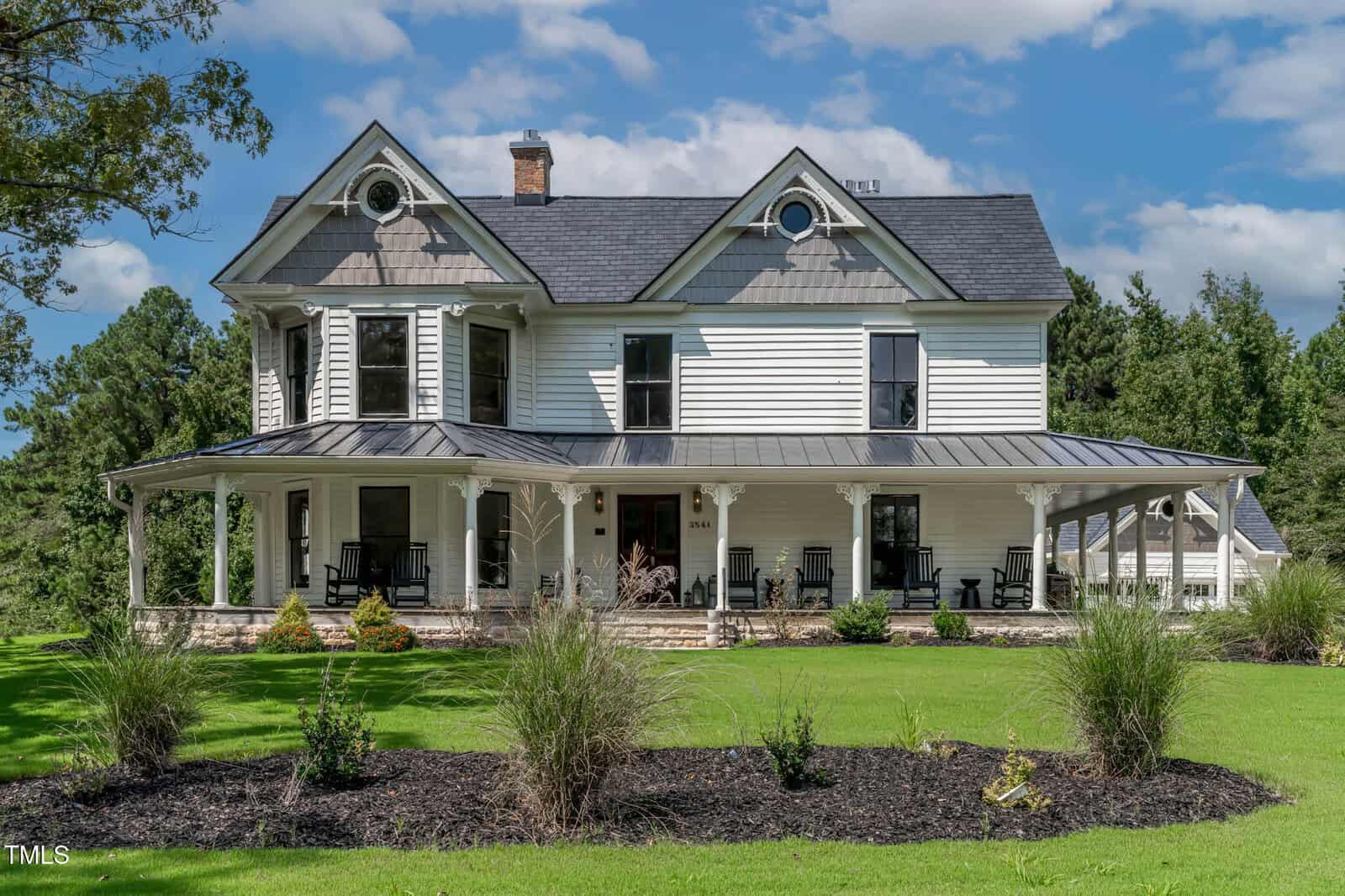 A large, white two-story house with a wraparound porch, black roof, and decorative gables, surrounded by green lawn, shrubs, and trees under a partly cloudy sky.