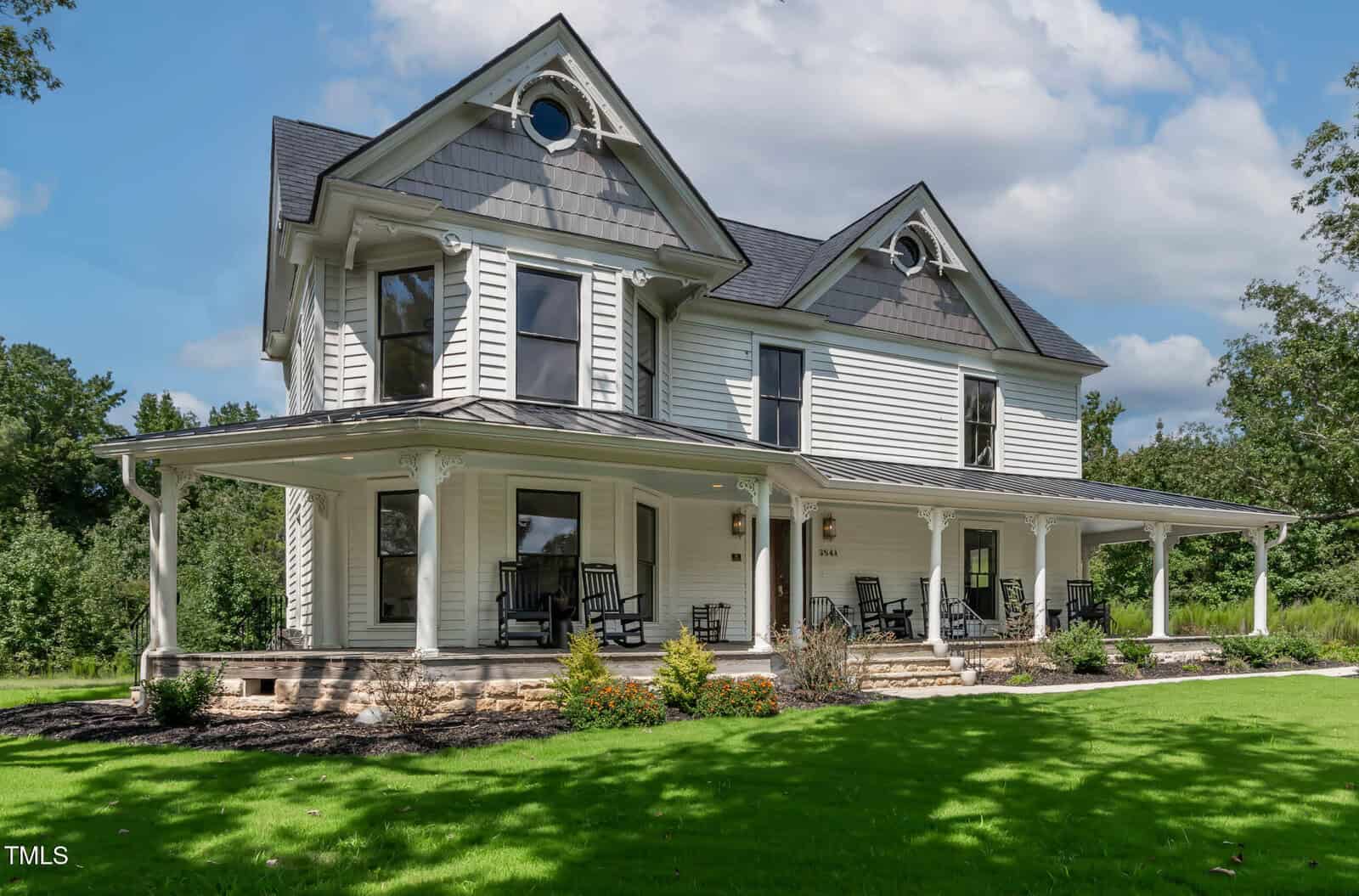 A large, white, two-story Victorian-style house with a wraparound porch, black rocking chairs, a manicured lawn, and trees in the background under a partly cloudy sky.