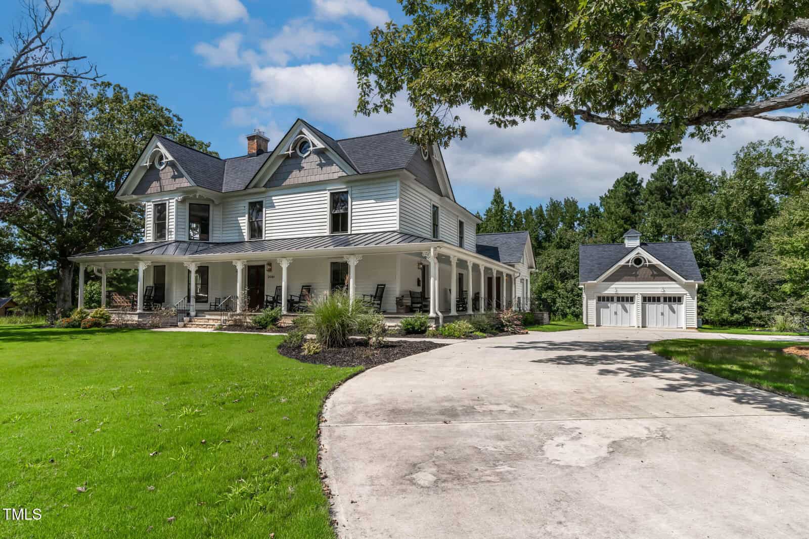 A large white two-story house with a wraparound porch sits beside a detached two-car garage. The home has dark gray roofs, decorative trim, and is surrounded by green lawn and trees under a blue sky.