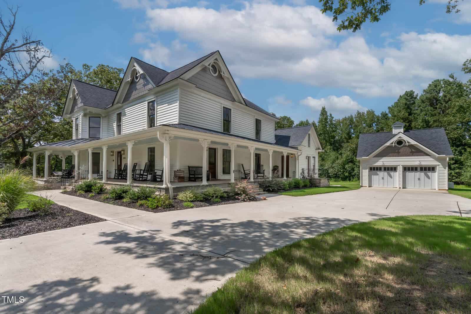 White two-story house with a wraparound porch, black roof, and rocking chairs, set on a spacious driveway with a detached two-car garage, surrounded by green lawn and trees under a partly cloudy sky.