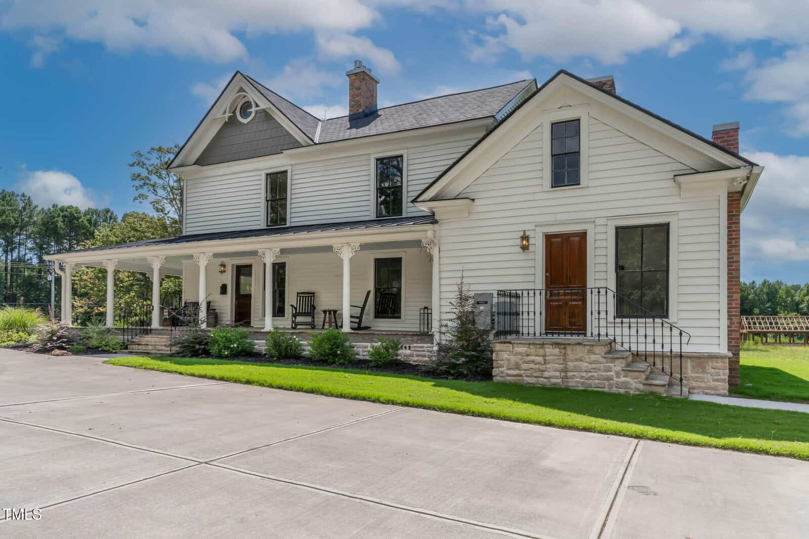 A large white two-story house with a front porch, wooden double doors, tall windows, rocking chairs, stone foundation, and a neatly trimmed lawn under a blue sky with scattered clouds.