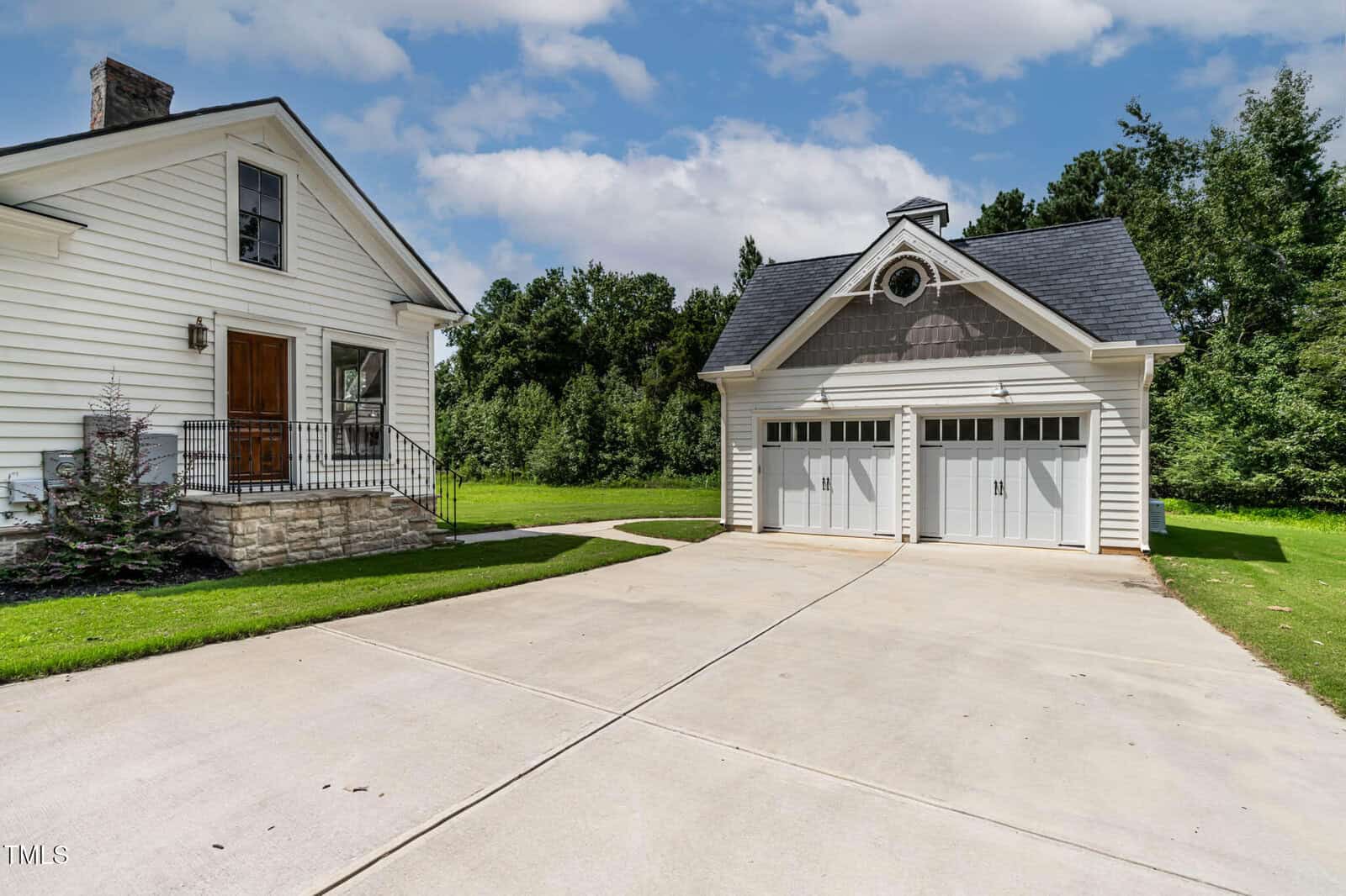 White two-story house with stone steps and a wooden door, next to a detached two-car garage. Concrete driveway in front, green lawn, and trees in the background under a partly cloudy sky.