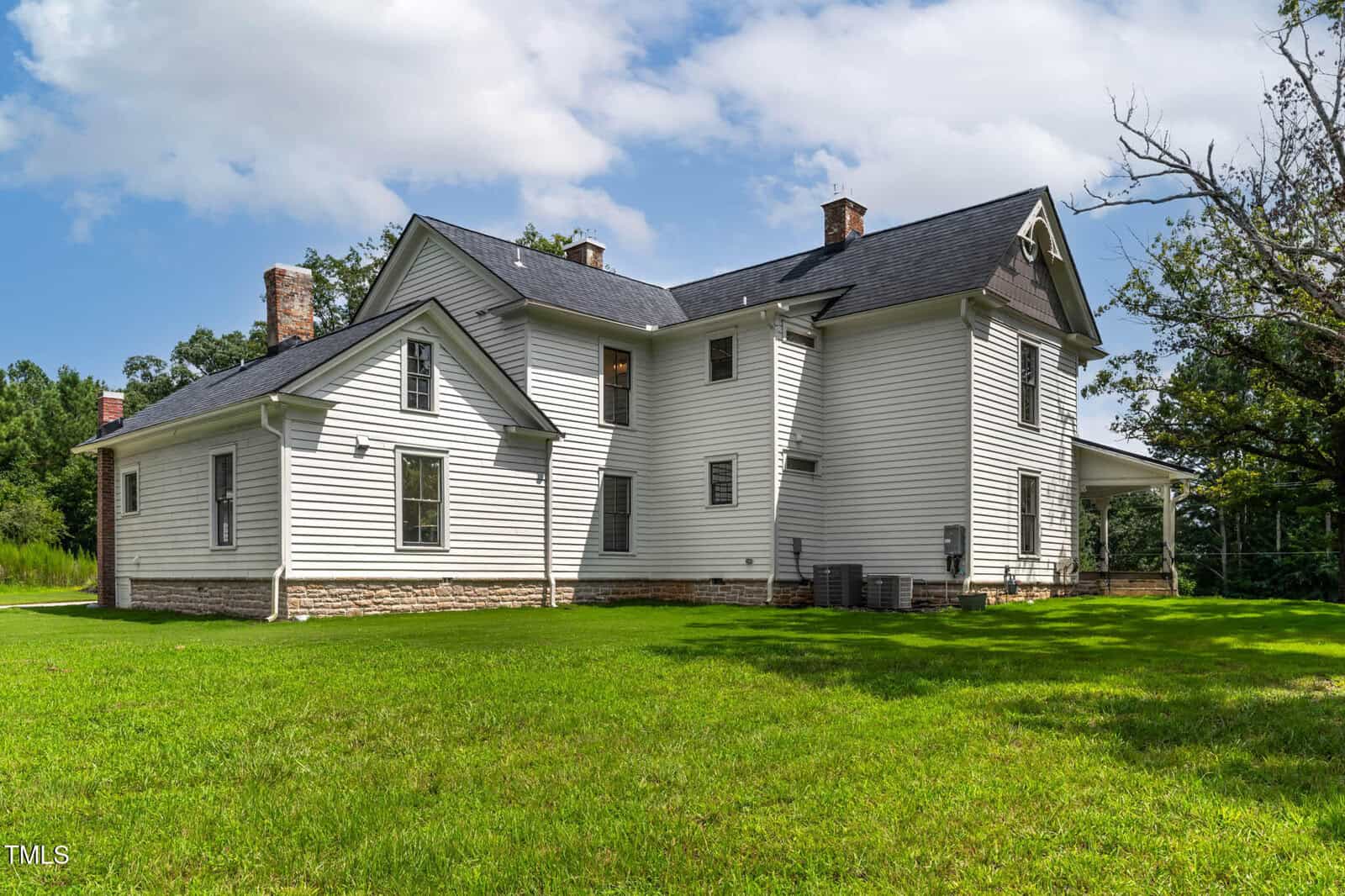 A white two-story house with a dark roof, several chimneys, and a wraparound porch, sits on a green lawn with trees and a blue sky with clouds in the background.