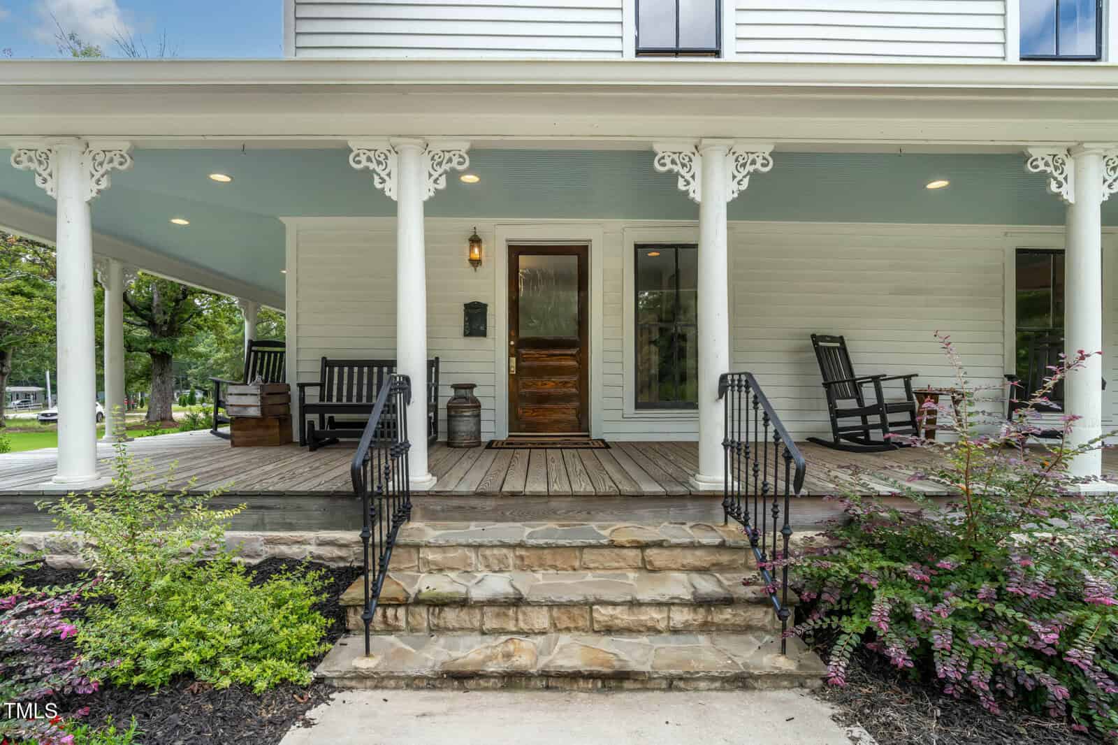 A white house porch with stone steps, white columns, black rocking chairs, a wooden front door, and decorative trim, set among green landscaping and trees.