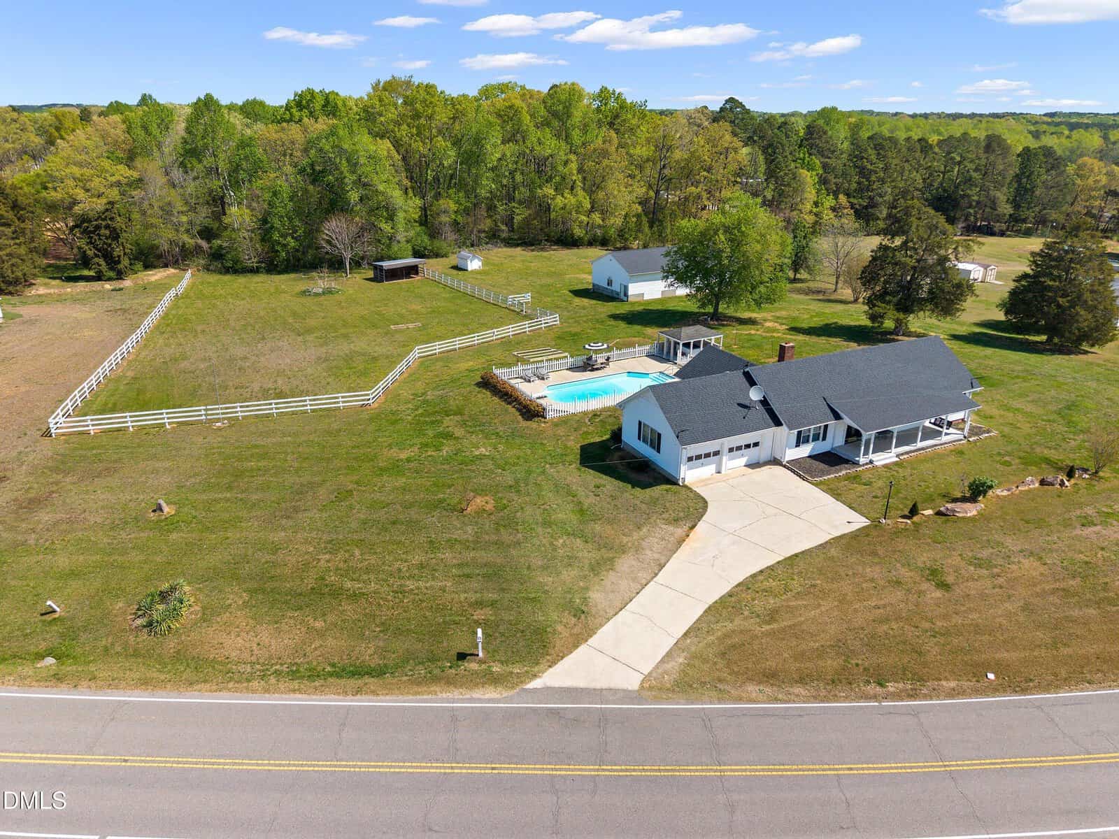 Aerial view of a rural property featuring a single-story house with a porch, a swimming pool, fenced yard, separate garage, and a large grassy area surrounded by trees, adjacent to a road.