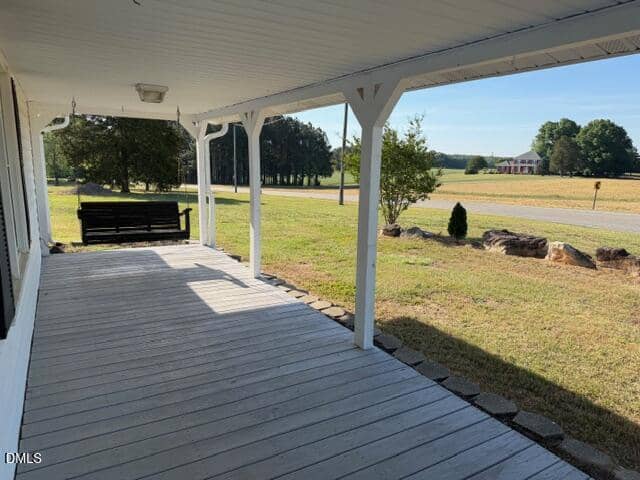 A wide, covered front porch with white columns overlooks a grassy yard and a country road. A black porch swing hangs at one end, and trees and a distant house are visible in the background.