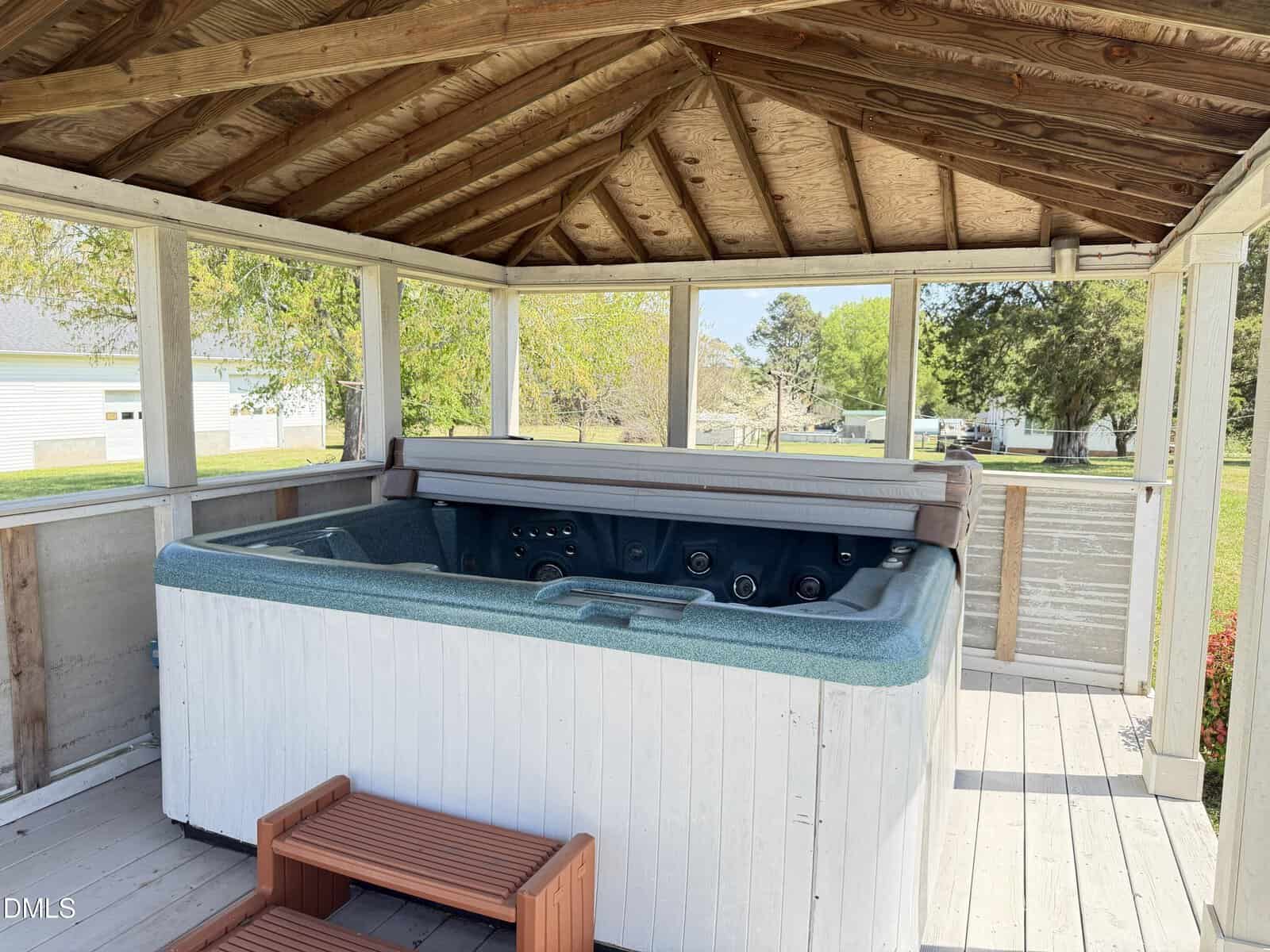 A covered outdoor hot tub with a blue interior and white exterior sits on a wooden deck. The hot tub cover is partially open, and there’s a small brown step in front. The area is surrounded by open panels and greenery outside.