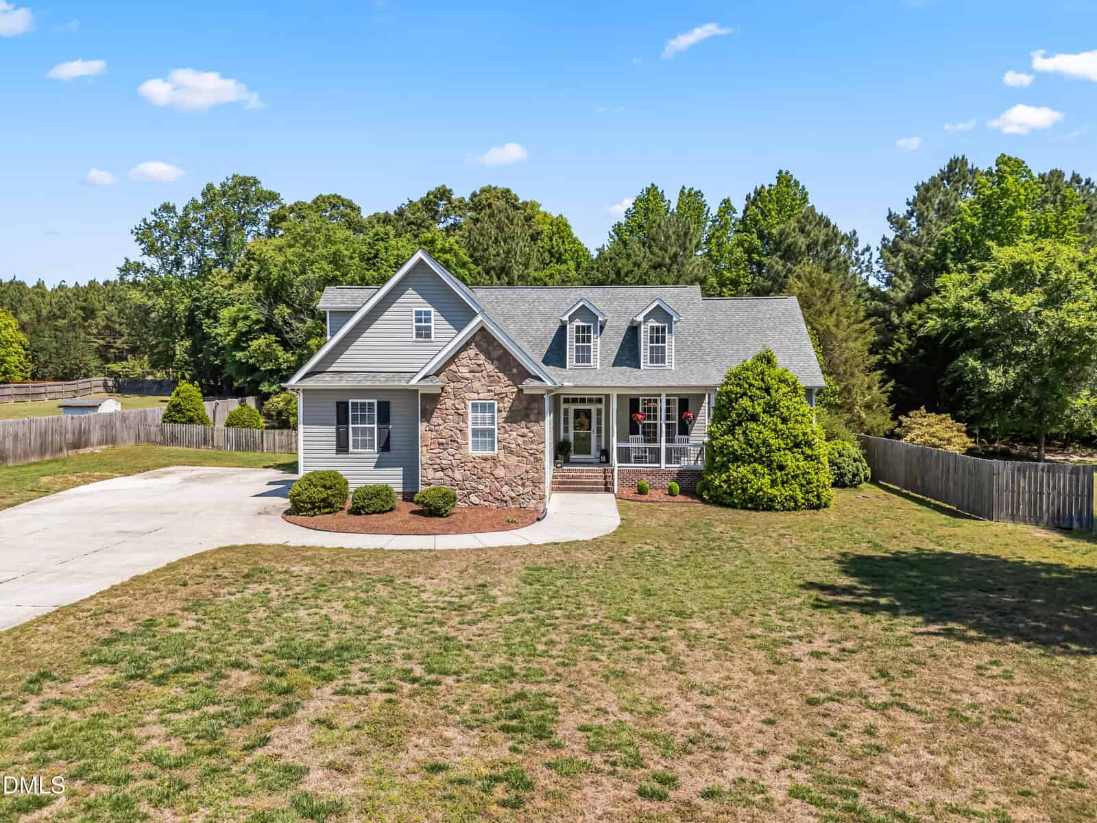 Two-story suburban house with stone and siding exterior, a front porch, and dormer windows. The home is surrounded by trees, has a large driveway, and sits on a spacious, partly grassy yard under a blue sky.