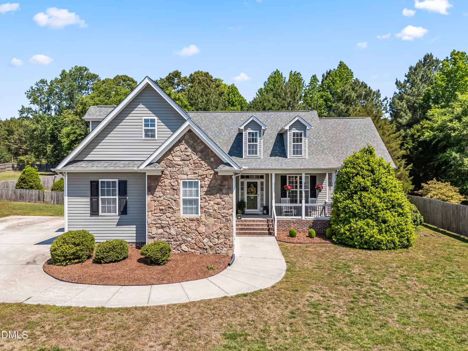 A gray house with stone accents, a front porch with red flowers, and a well-maintained lawn, sits under a blue sky with scattered clouds, surrounded by trees and a curved driveway.