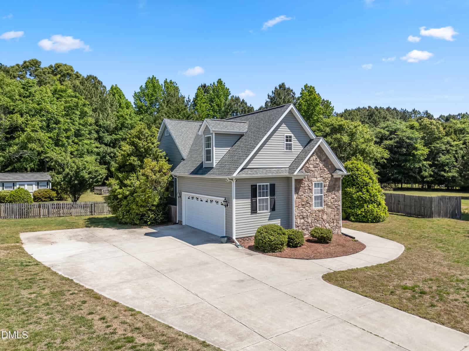 A two-story house with gray siding and stone accents is surrounded by trees, featuring a curved driveway, attached garage, and well-kept front landscaping under a clear blue sky.