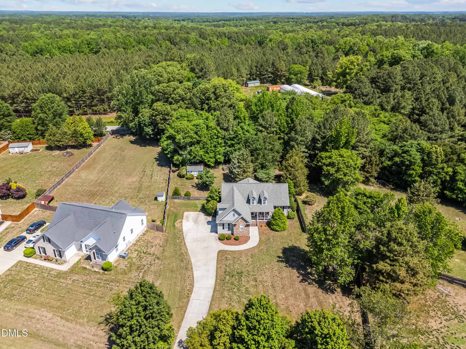 Aerial view of a grey house with a curved driveway, surrounded by trees and greenery, with a neighboring white house and spacious lawns in a rural area. Dense forest stretches in the background.