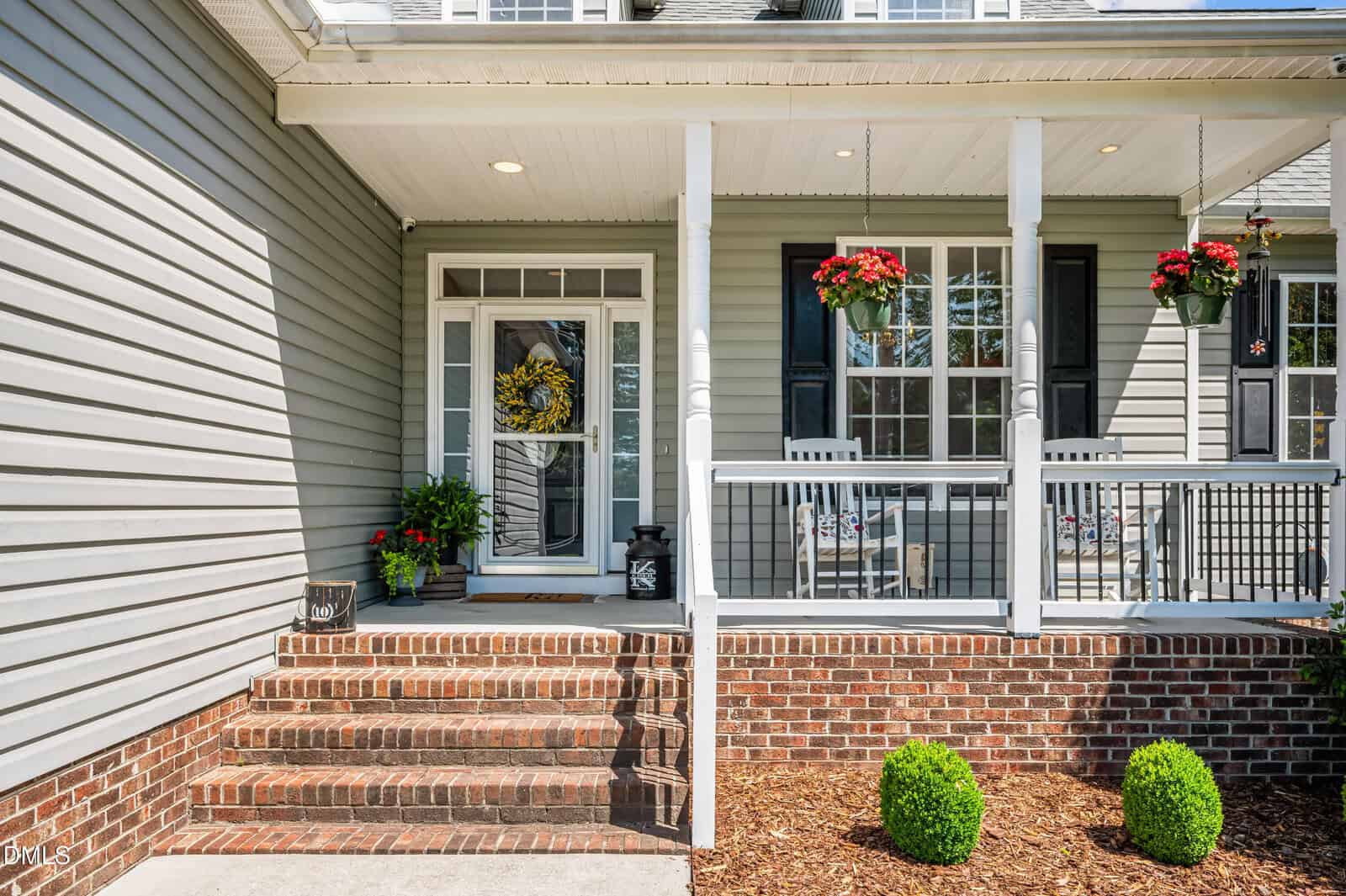 A brick staircase leads to a covered front porch with gray siding. There are white railings, hanging flower baskets, a welcome sign, a potted plant, a yellow wreath on the front door, and two trimmed shrubs by the steps.
