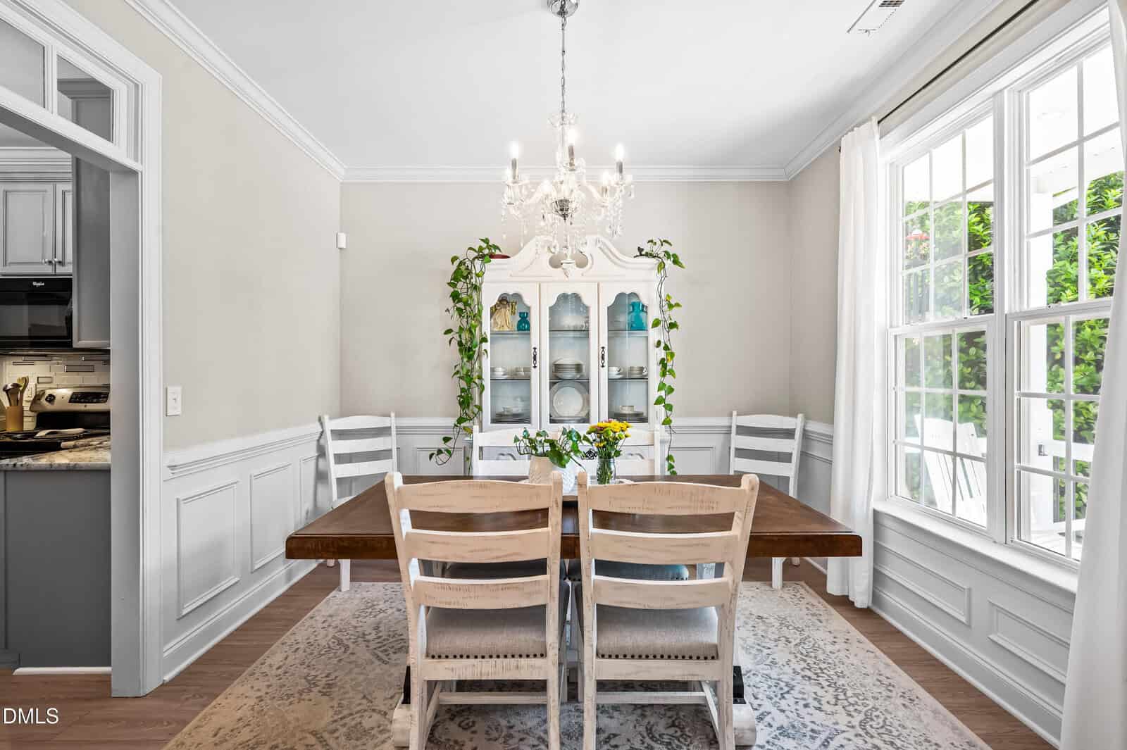 Bright dining room with a wooden table, four white chairs, chandelier, china cabinet with plants, and large window letting in natural light.