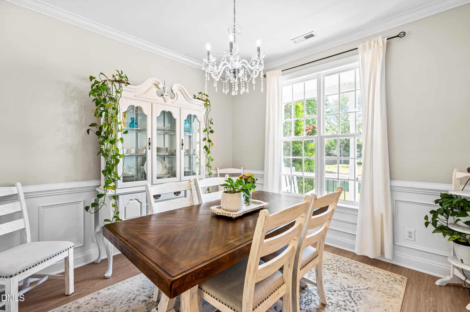 Bright dining room with a wooden table, four white chairs, a chandelier, a china cabinet, potted plants, and a large window with white curtains letting in natural light.