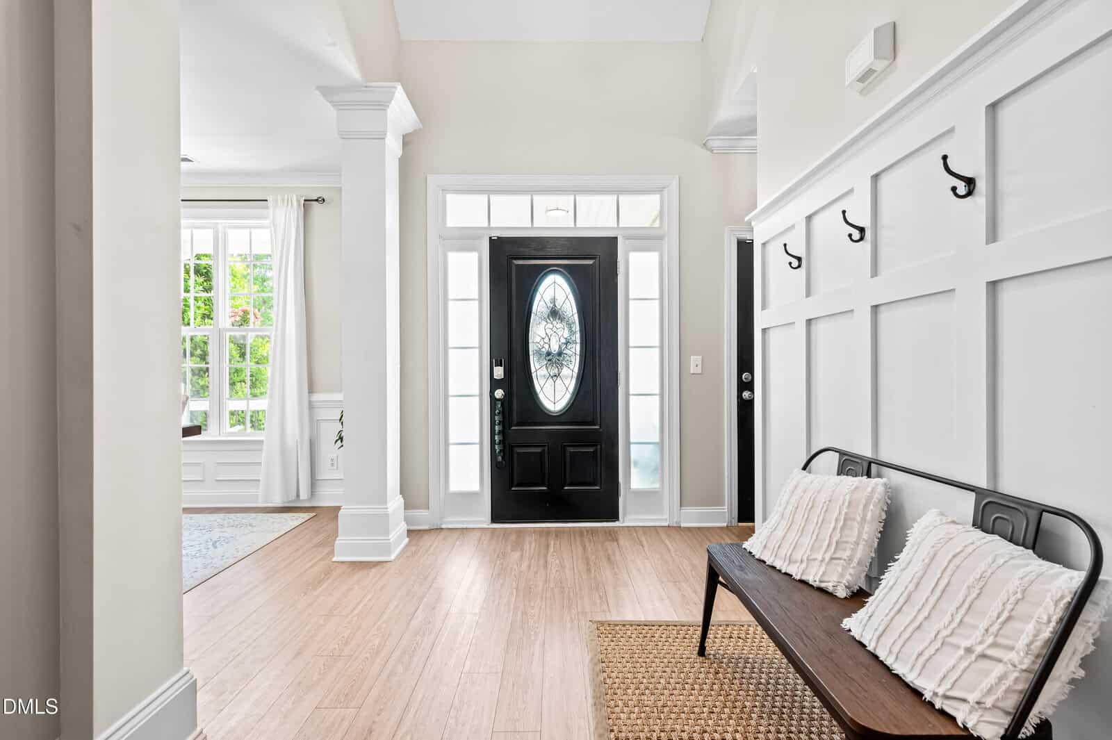 Bright entryway with a black front door, side and transom windows, a wooden bench with cream pillows, wall hooks, white walls, and light wood flooring. Sunlight streams through large windows on the left.