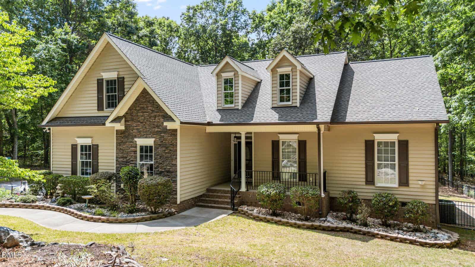 A beige two-story house with stone accents, dormer windows, and a covered front porch, surrounded by shrubs and trees with a curved walkway leading to the front door.