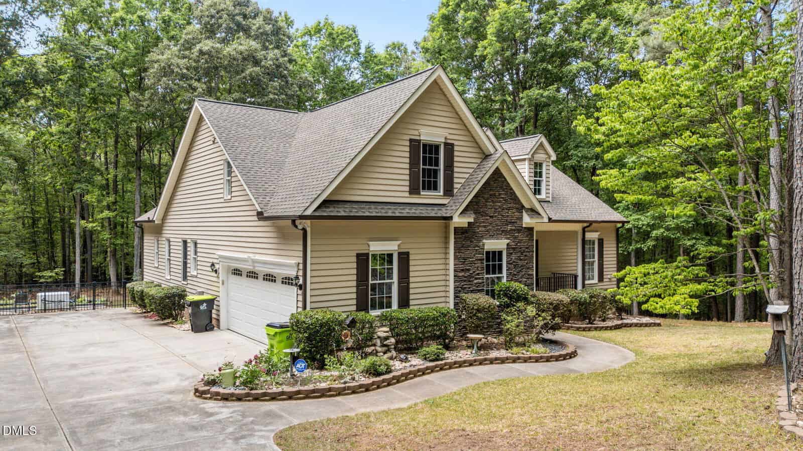 A tan two-story house with stone accents, a gabled roof, white trim, and a three-car garage sits among tall trees. A curved driveway, trimmed bushes, and landscaped flower beds enhance the front yard.
