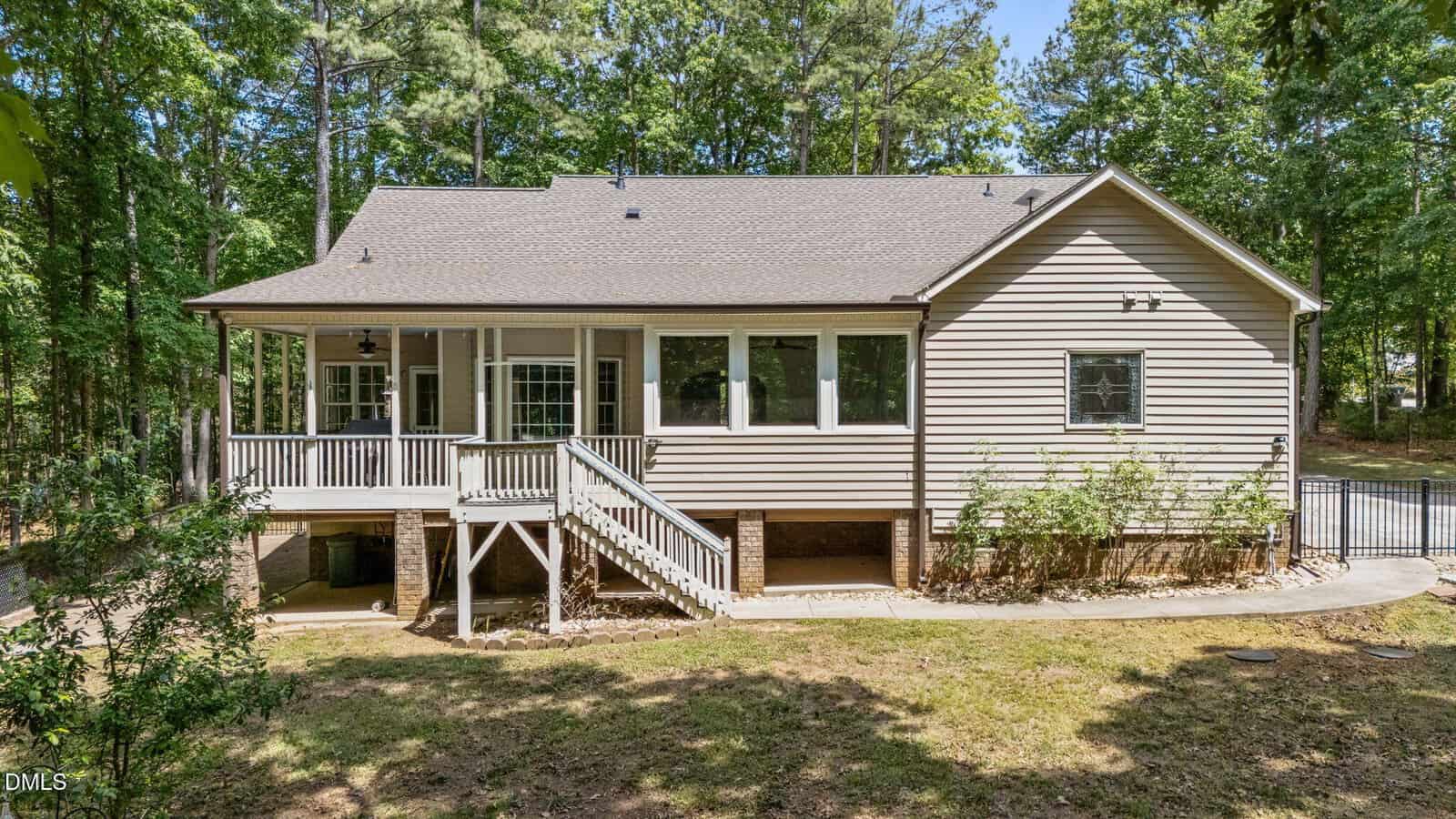 A light brown, single-story house with a covered porch and white railings, surrounded by trees. Wooden steps lead to the yard, and there are brick supports and shrubbery along the foundation.