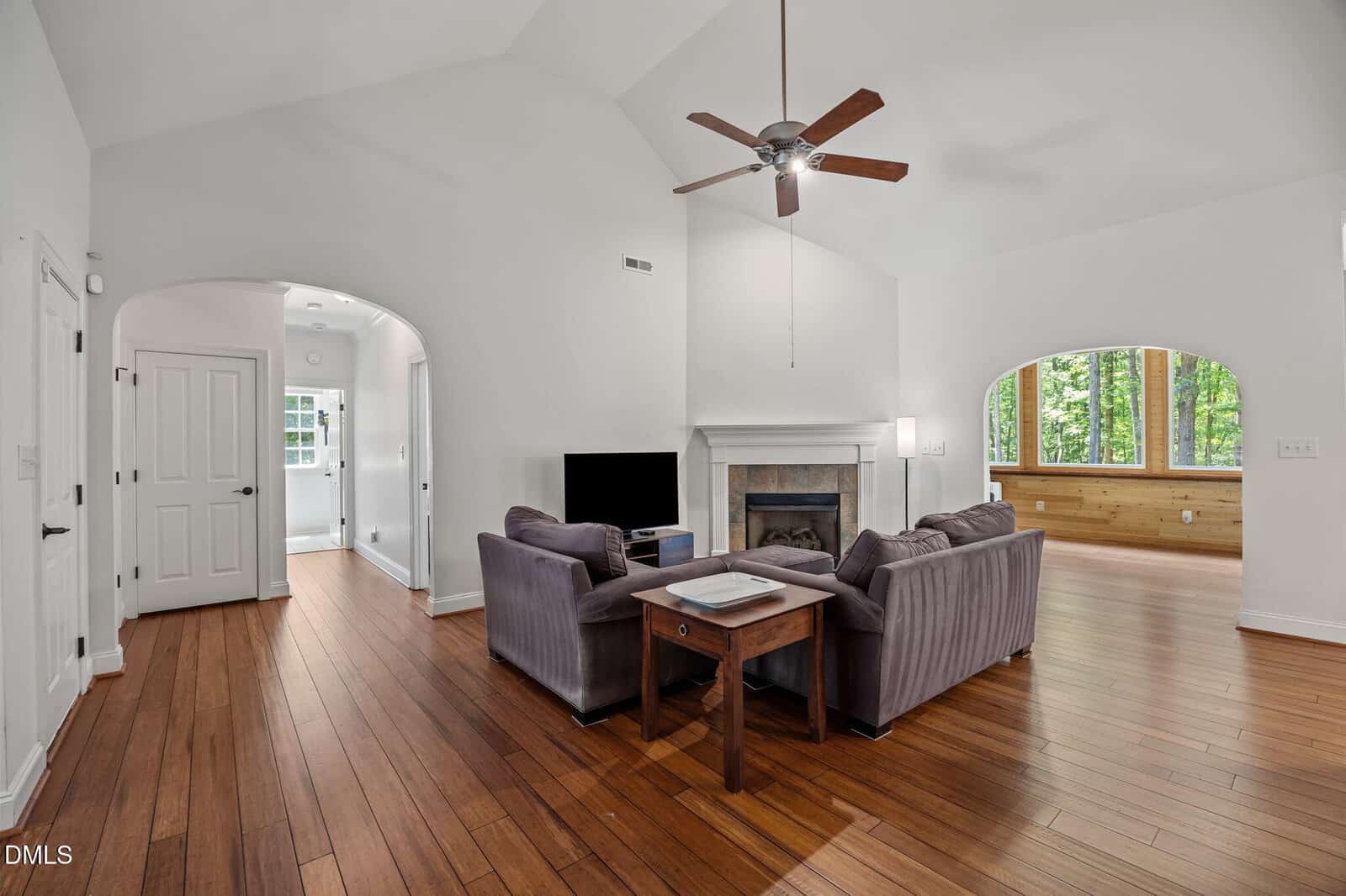 Bright living room with vaulted ceiling, ceiling fan, hardwood floors, two gray sofas, a fireplace, TV, and large windows overlooking a sunroom with wooded views. White walls and doorways add to the open, airy feel.
