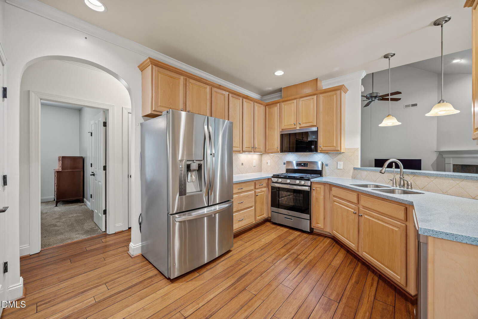 Modern kitchen with light wood cabinets, stainless steel appliances, gas stove, double sink, and wood flooring. The space is open, bright, and includes an arched doorway leading to another room.