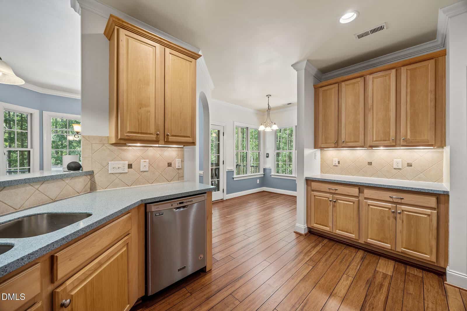 A spacious kitchen with light wood cabinets, stainless steel dishwasher, light countertops, tile backsplash, and hardwood floors, opening to a bright dining area with large windows and a view of greenery outside.