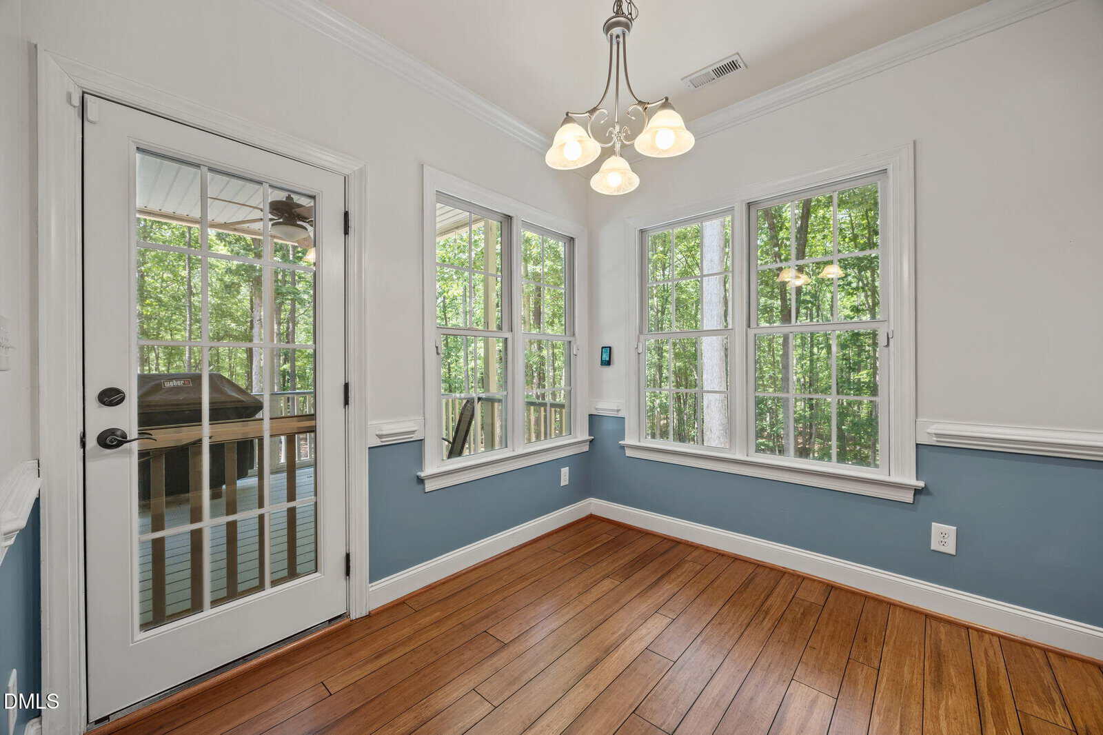 A small dining area with wood flooring, blue and white walls, large windows, a glass door leading to a screened porch with a grill, and a ceiling light fixture. Trees are visible outside the windows.